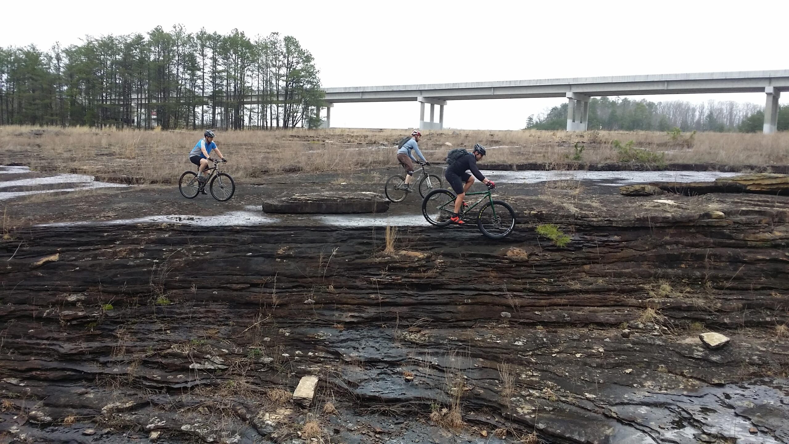 Three mountain bikers riding on rocky terrain with sparse vegetation, under a cloudy sky. In the background, there is a highway bridge and a line of trees. The landscape appears rugged and natural, indicating an outdoor biking adventure. Sheltowee Trace - Laurel Lake Trail mountain bike trail.