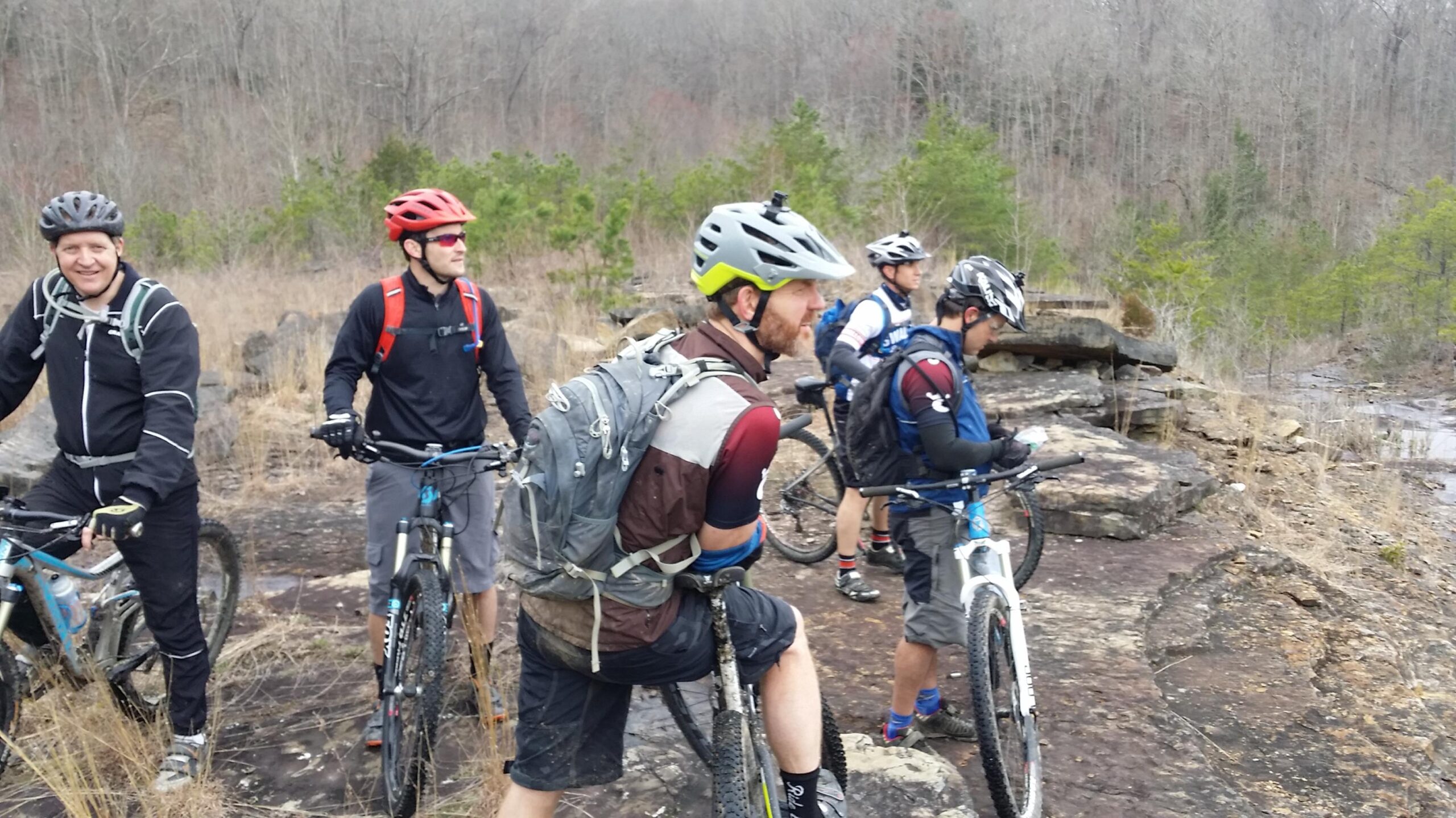 A group of five mountain bikers atop a rocky outcrop, wearing helmets and cycling gear. They are surrounded by sparse vegetation and trees in the background, with a hint of a river or stream nearby. The bikers are discussing or observing the scenery, and their mountain bikes are parked on the rocky surface. Sheltowee Trace - Laurel Lake Trail mountain bike trail.