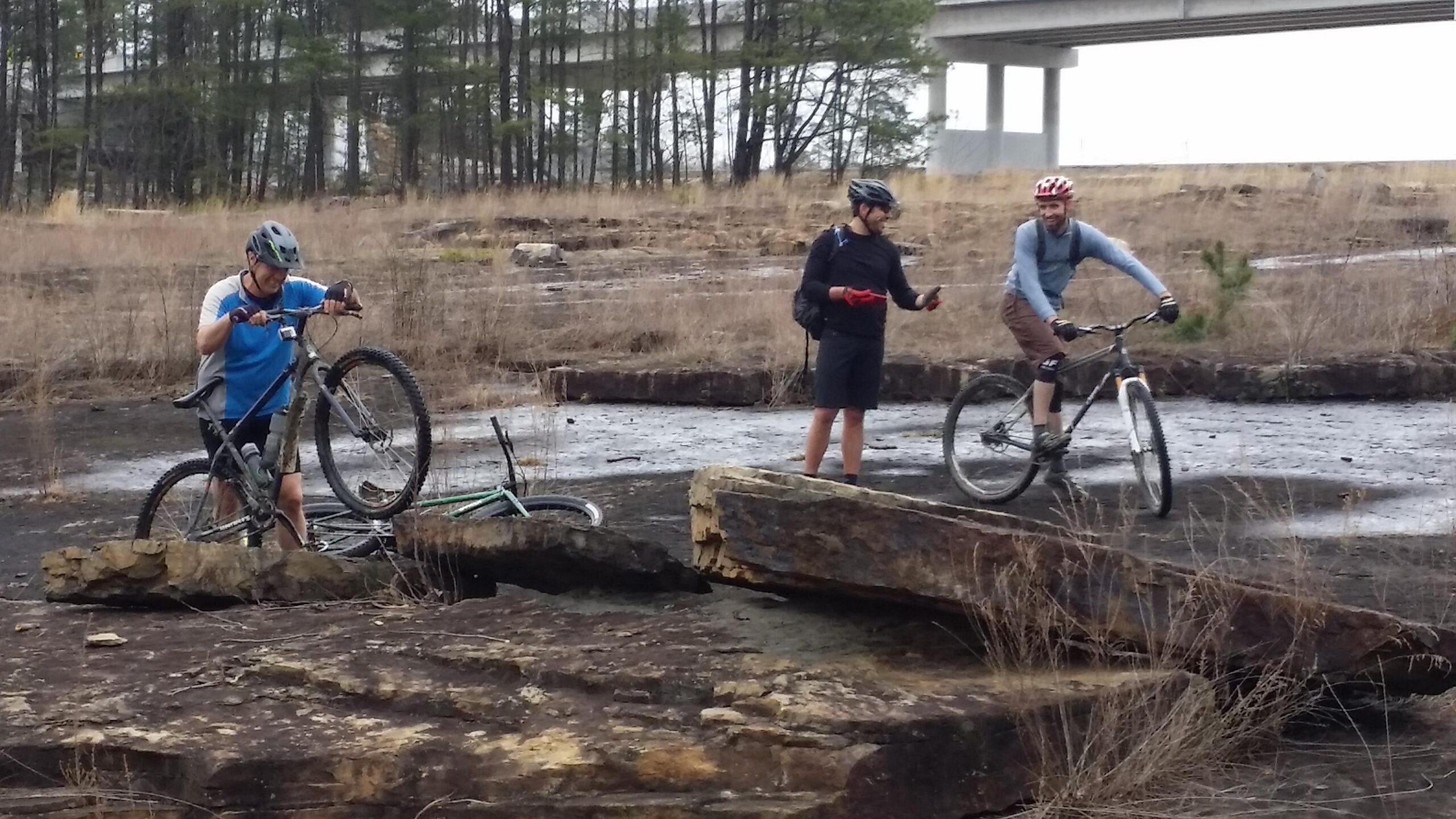 Three mountain bikers are outdoors on rocky terrain, with patches of dry grass in the background. One rider, in a blue shirt and helmet, is adjusting his bike, lifting the front wheel off the ground. Another, wearing a black shirt and holding a phone, stands nearby, while a third rider, dressed in a light blue top and brown shorts, is riding his bike across the uneven surface. In the background, a highway overpass is visible among trees. Sheltowee Trace - Laurel Lake Trail mountain bike trail.