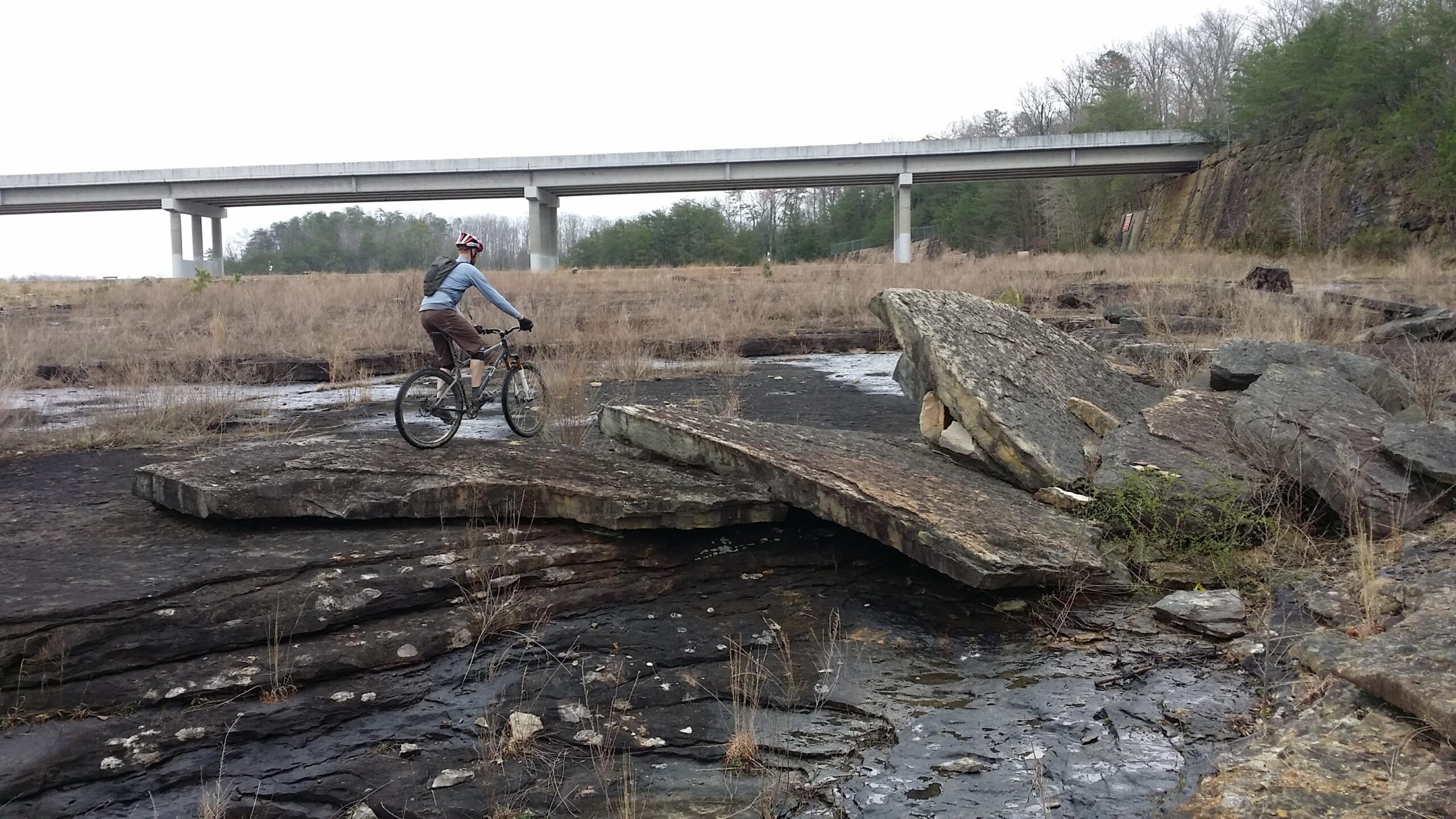 A mountain biker rides on a large rock formation in a grassy area, with a concrete overpass visible in the background. The scene features rocky terrain with sparse vegetation and a cloudy sky above. Sheltowee Trace - Laurel Lake Trail mountain bike trail.