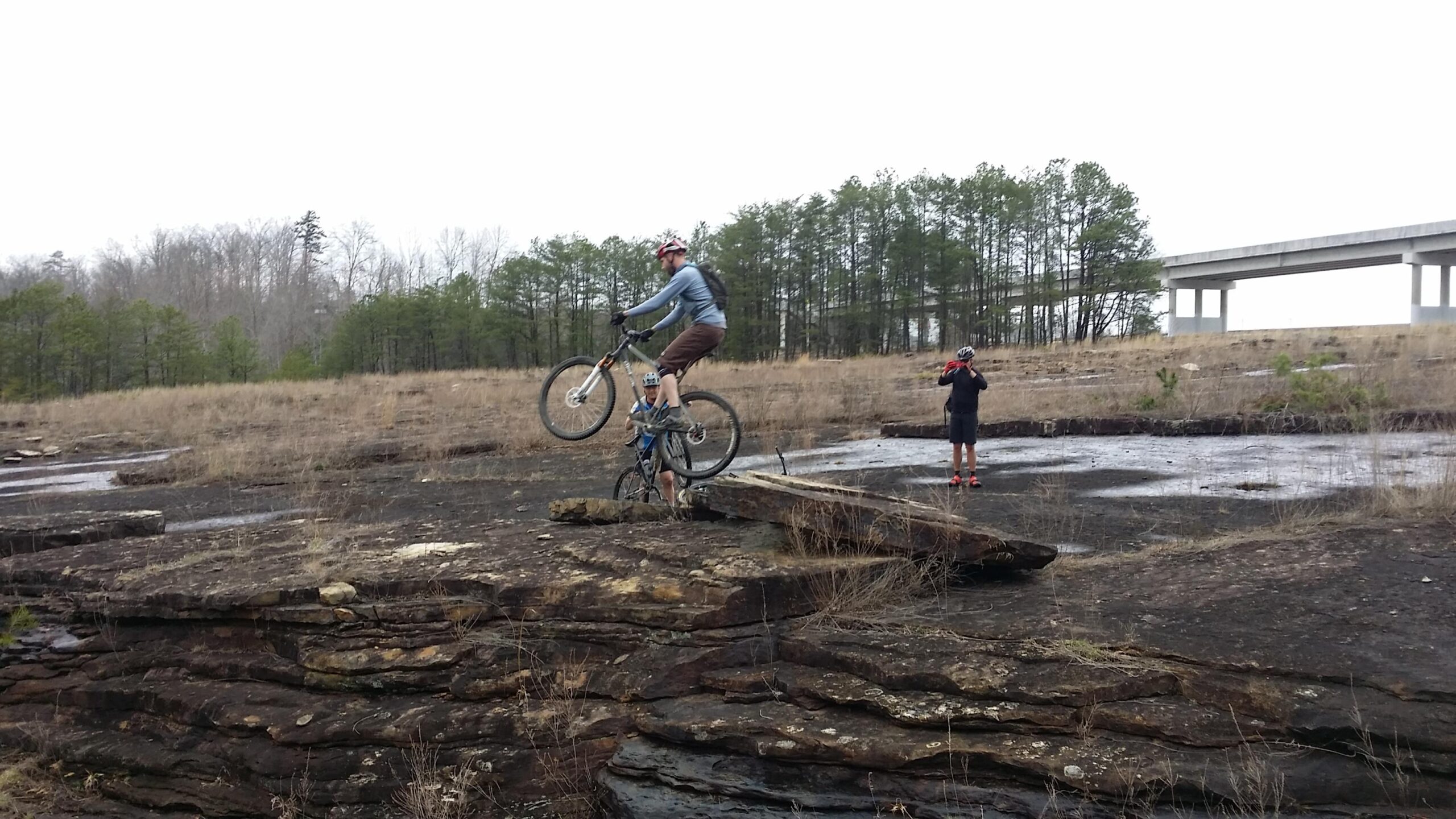 A mountain biker performing a jump over a rocky ledge while another cyclist watches nearby. In the background, a grassy landscape and a bridge are visible against a cloudy sky. Sheltowee Trace - Laurel Lake Trail mountain bike trail.