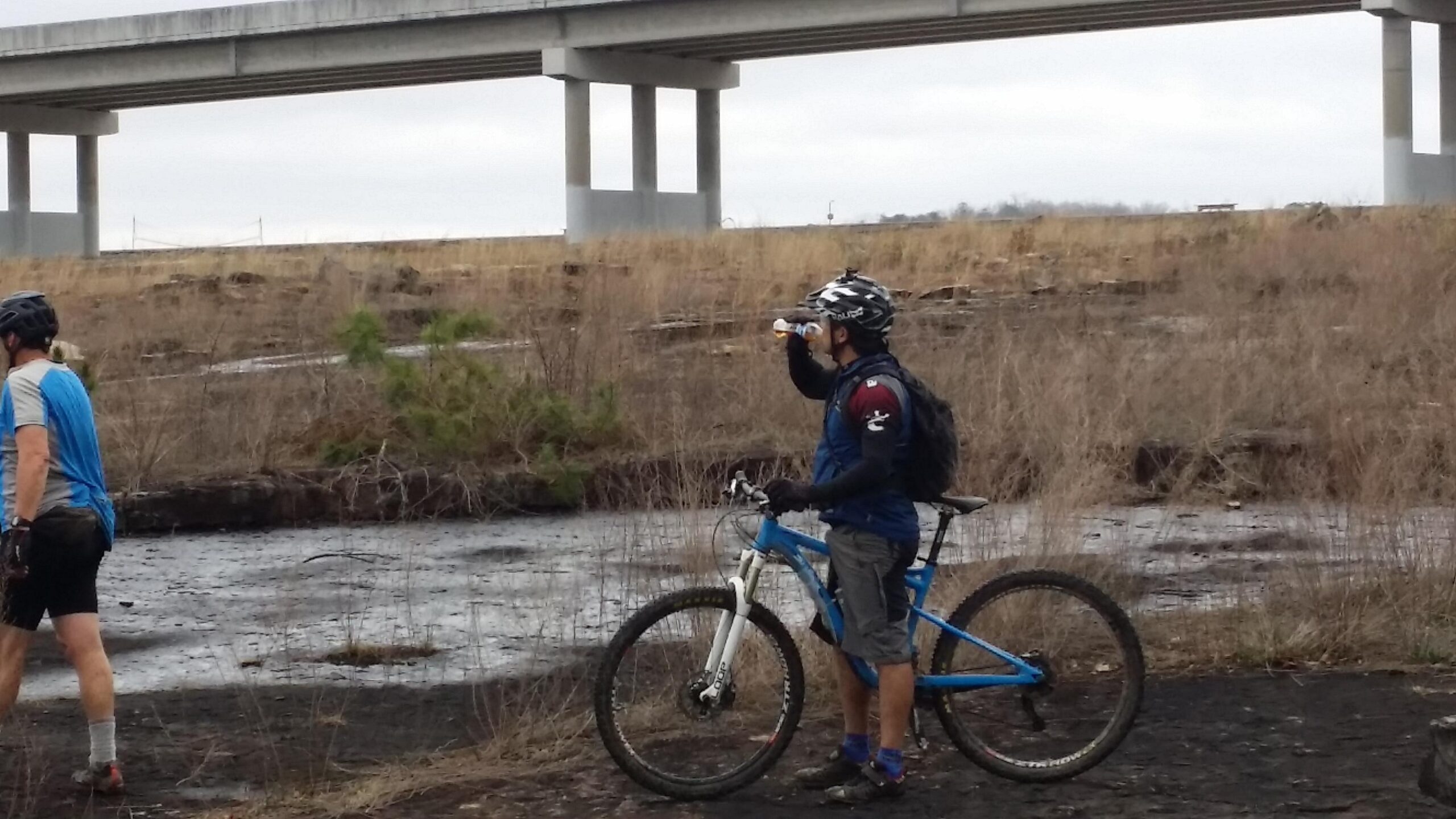 A mountain biker pauses to drink water while standing next to his blue bicycle in a grassy area. In the background, a concrete overpass is visible, with bare trees and a muddy ground in the foreground, indicating an outdoor biking trail. Sheltowee Trace - Laurel Lake Trail mountain bike trail.