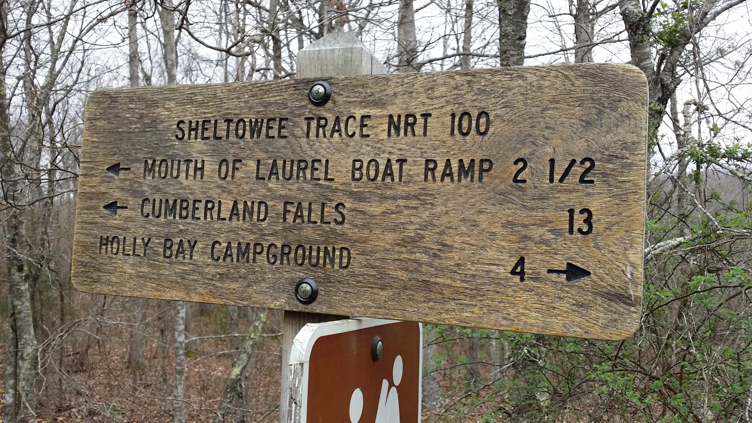 A weathered wooden sign displaying trail directions for the Sheltowee Trace National Recreational Trail (NRT 100). The sign indicates distances to the Mouth of Laurel Boat Ramp (2.5 miles), Cumberland Falls (13 miles), and Holly Bay Campground (4 miles), with directional arrows pointing left and right. The background features a forested area with bare trees and underbrush. Laural Lake To Cumberland Falls (sheltowee Trace) mountain bike trail.