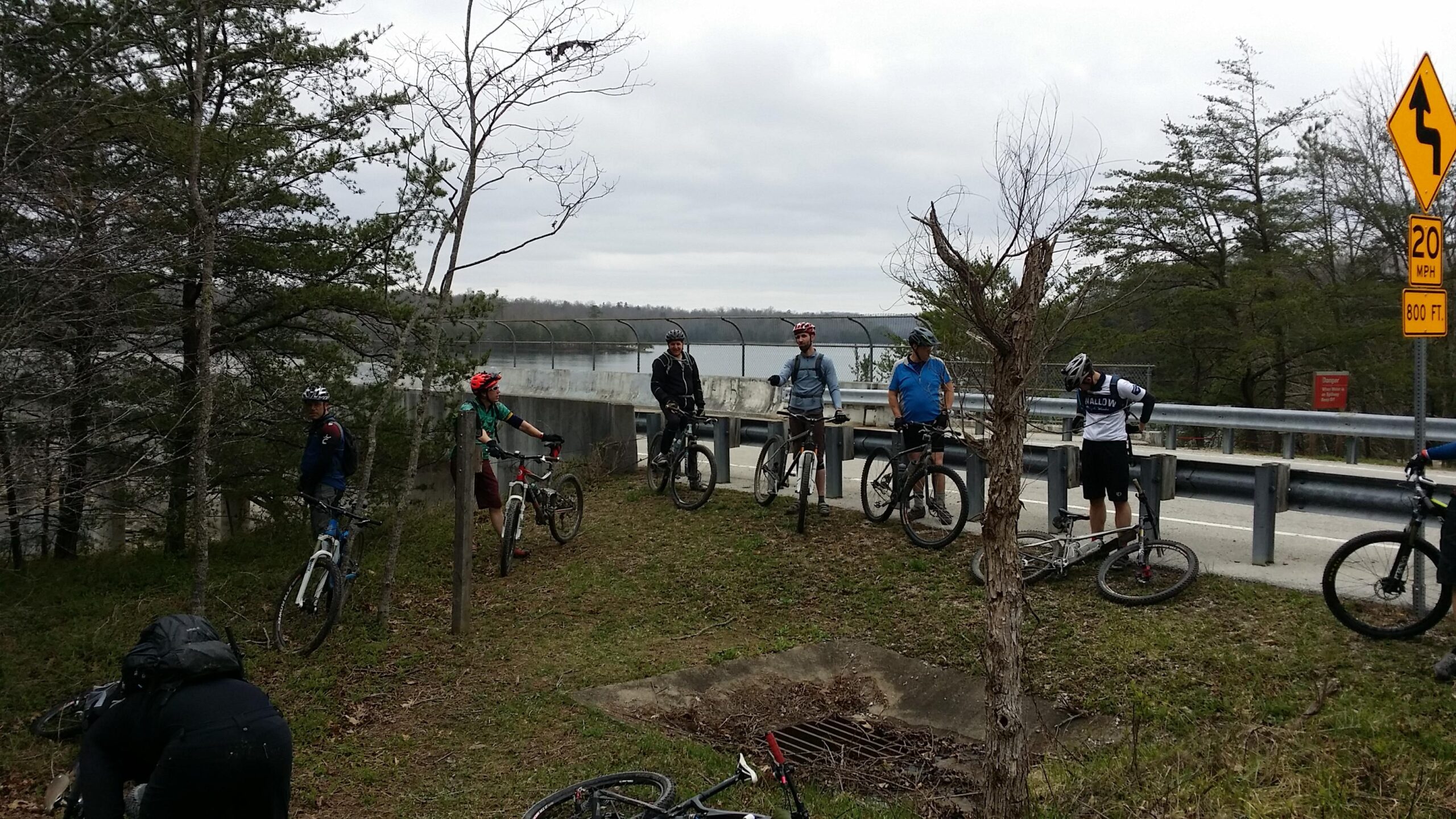 A group of five mountain bikers is gathered by a roadside near a body of water, surrounded by trees. They are wearing helmets and casual biking attire, with their bicycles parked nearby. The weather appears overcast. A warning sign indicating a turn is visible in the background. Laural Lake To Cumberland Falls (sheltowee Trace) mountain bike trail.