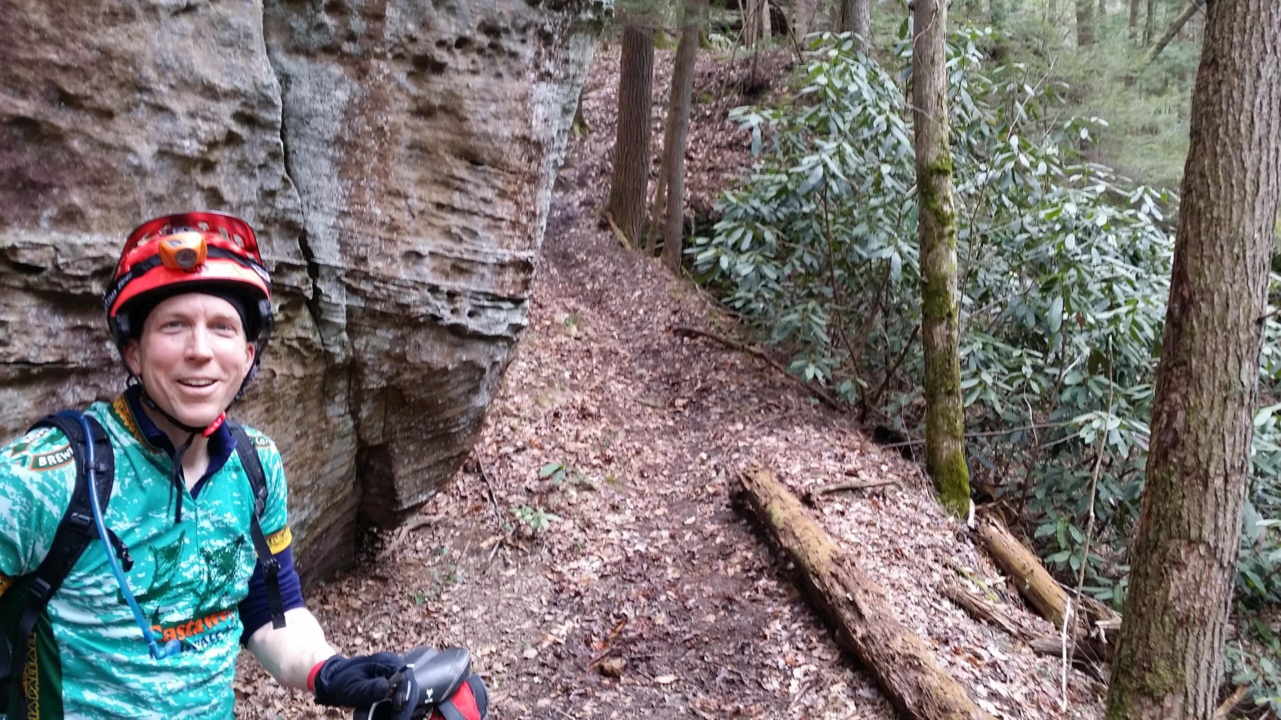 A person wearing a red helmet and a green cycling jersey stands in a forested area near a rocky outcrop. They are smiling and holding a cycling device. The background features trees and leafy underbrush, indicating a biking trail. Laural Lake To Cumberland Falls (sheltowee Trace) mountain bike trail.