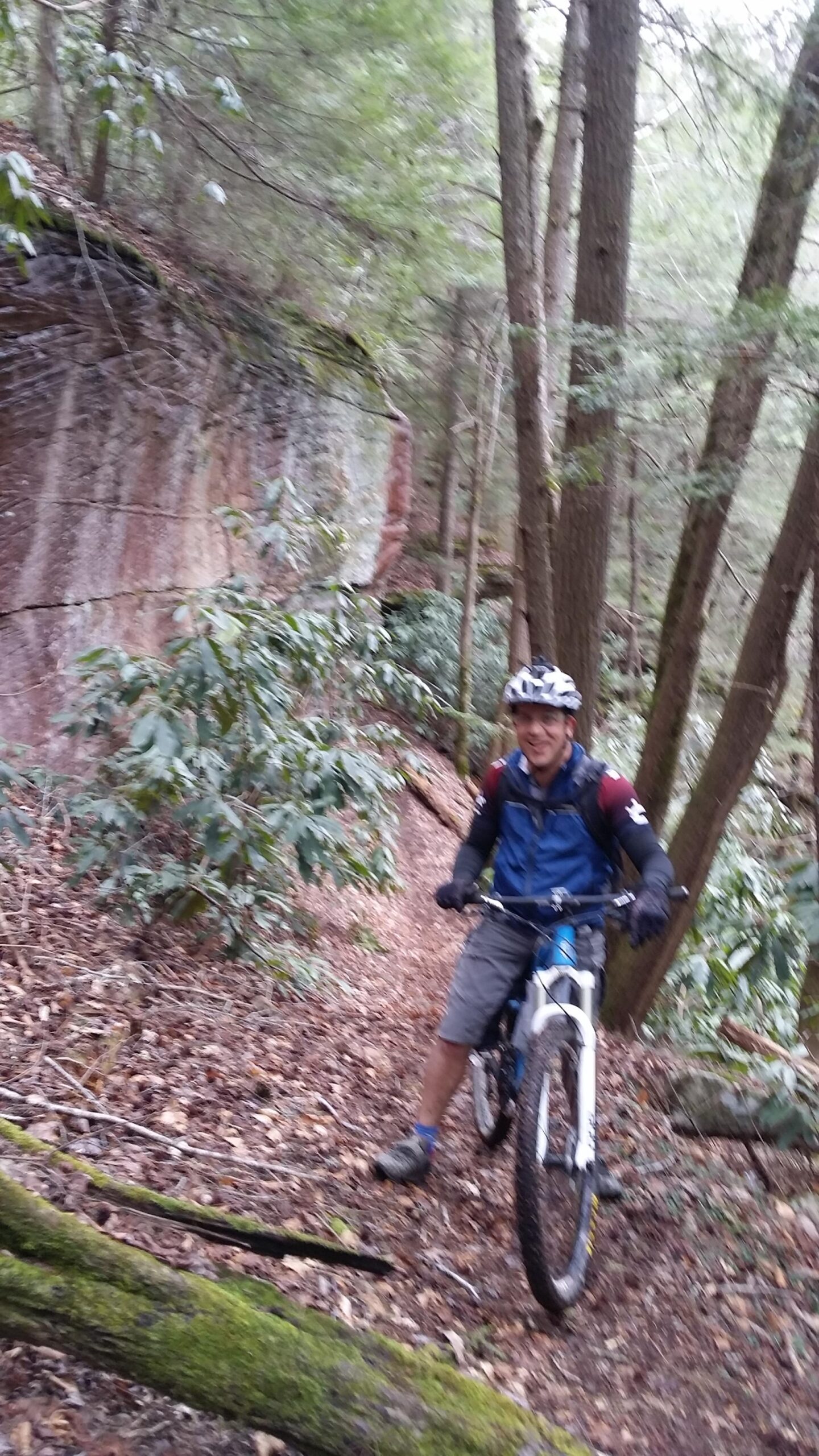 A person on a mountain bike navigating a narrow, wooded trail surrounded by trees and greenery, with a rocky cliff in the background. The cyclist is wearing a helmet, gloves, and a blue jacket, smiling while seated on the bike. Laural Lake To Cumberland Falls (sheltowee Trace) mountain bike trail.