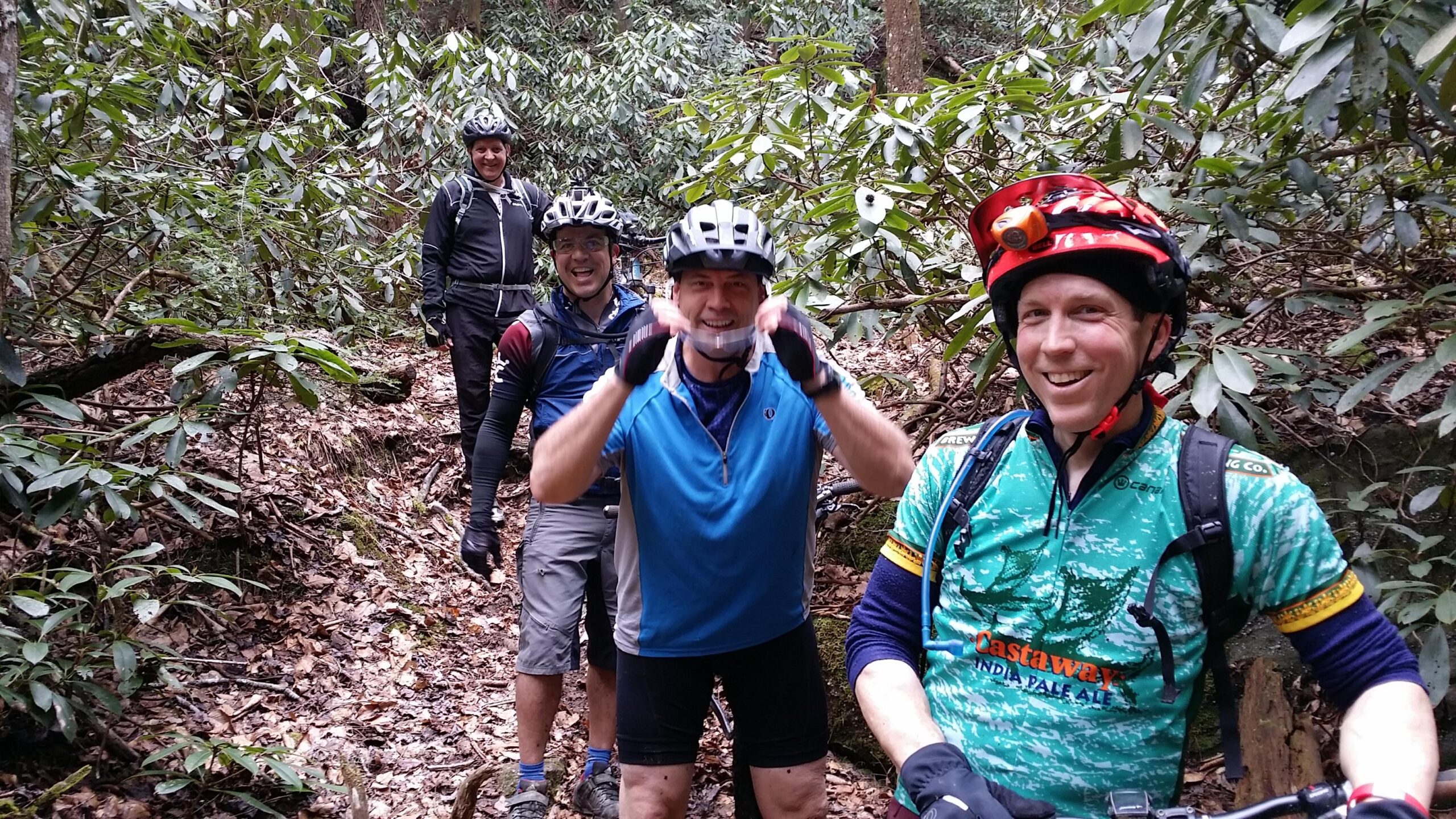 Four mountain bikers are smiling and posing for a photo on a wooded trail surrounded by lush green foliage. They are wearing helmets and cycling gear, with one biker showing a playful expression by raising his arms. The ground is covered with fallen leaves, and the atmosphere appears friendly and adventurous. Laural Lake To Cumberland Falls (sheltowee Trace) mountain bike trail.