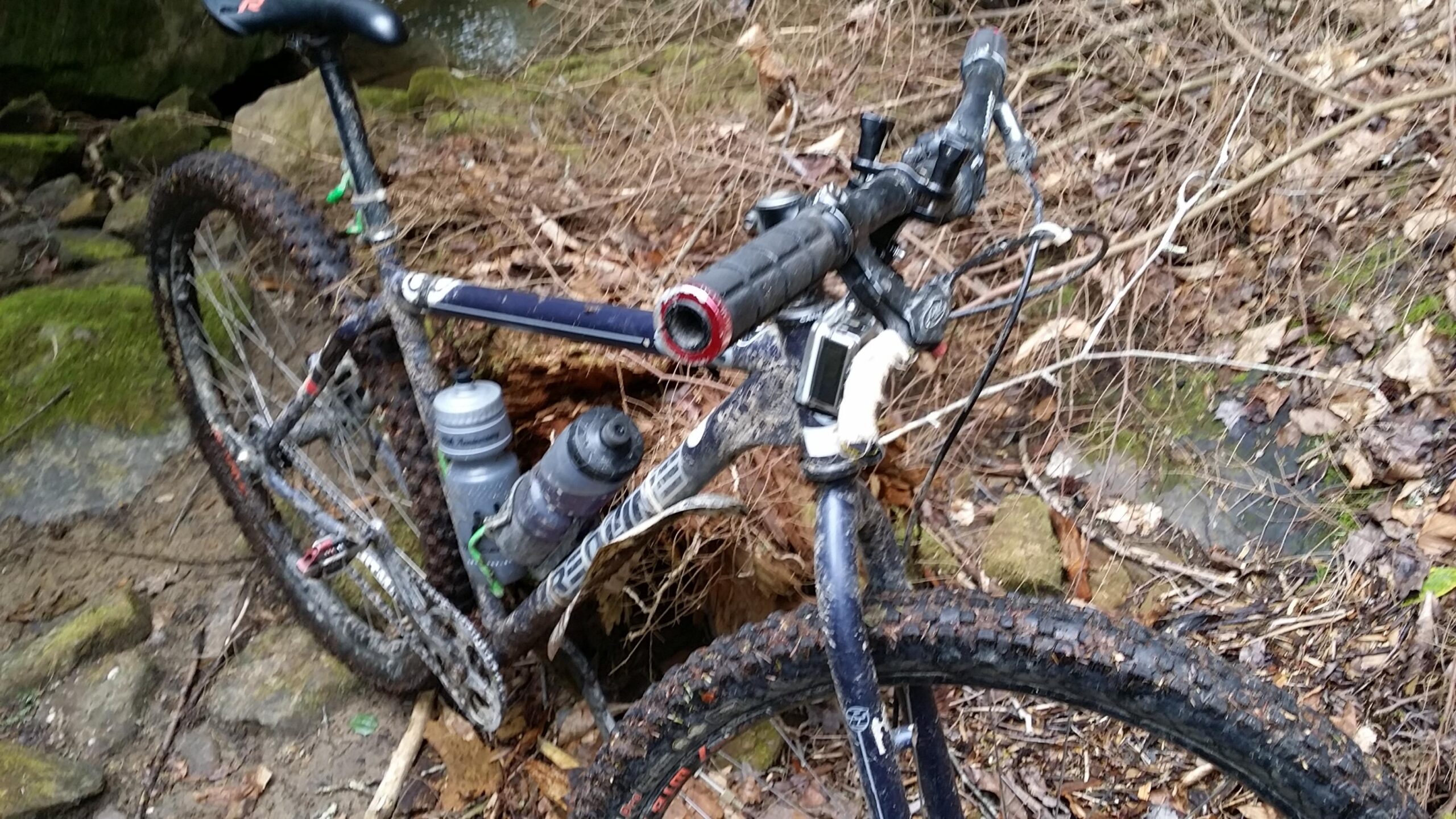 A mountain bike with muddy tires and dirt-covered frame is parked beside a rocky area, surrounded by fallen leaves and branches. A water bottle is attached to the frame, and the bike shows signs of recent use in off-road conditions. Laural Lake To Cumberland Falls (sheltowee Trace) mountain bike trail.