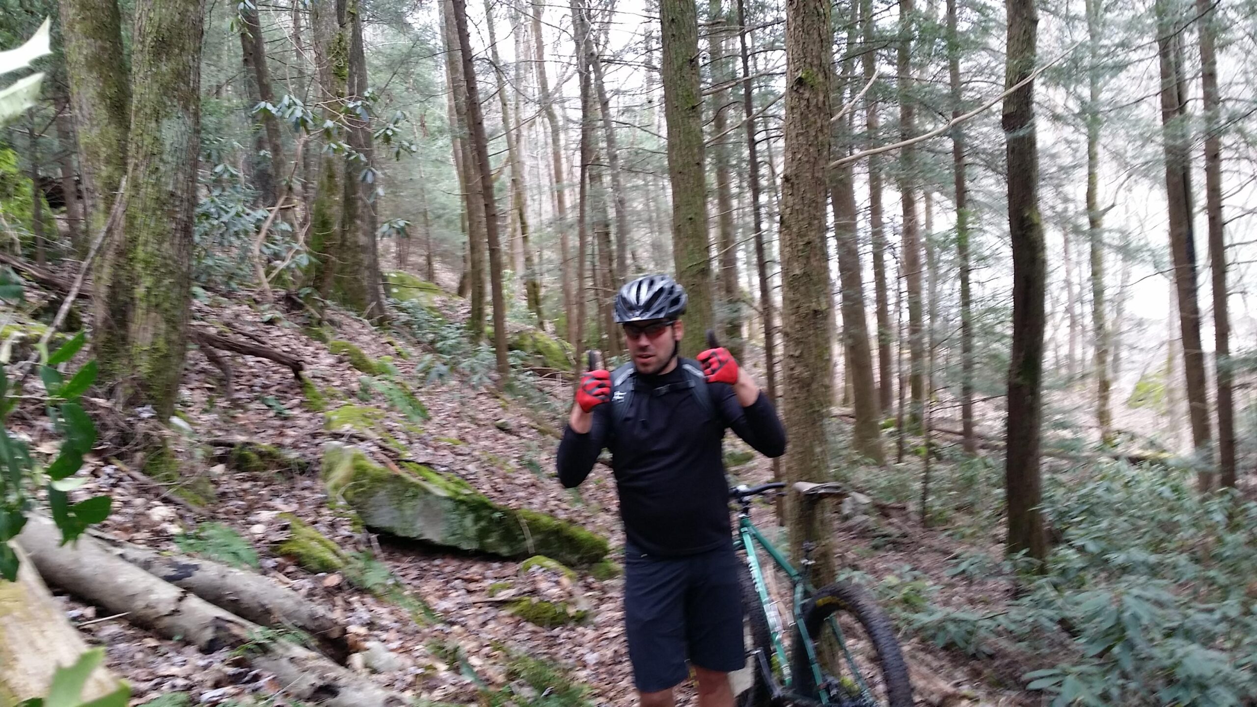 A mountain biker wearing a black helmet, black shirt, and red gloves is giving a thumbs-up gesture while standing on a forest trail. Behind him, there are tall trees and lush greenery, with a bicycle nearby. The setting is rocky and wooded, indicating a natural outdoor environment. Laural Lake To Cumberland Falls (sheltowee Trace) mountain bike trail.