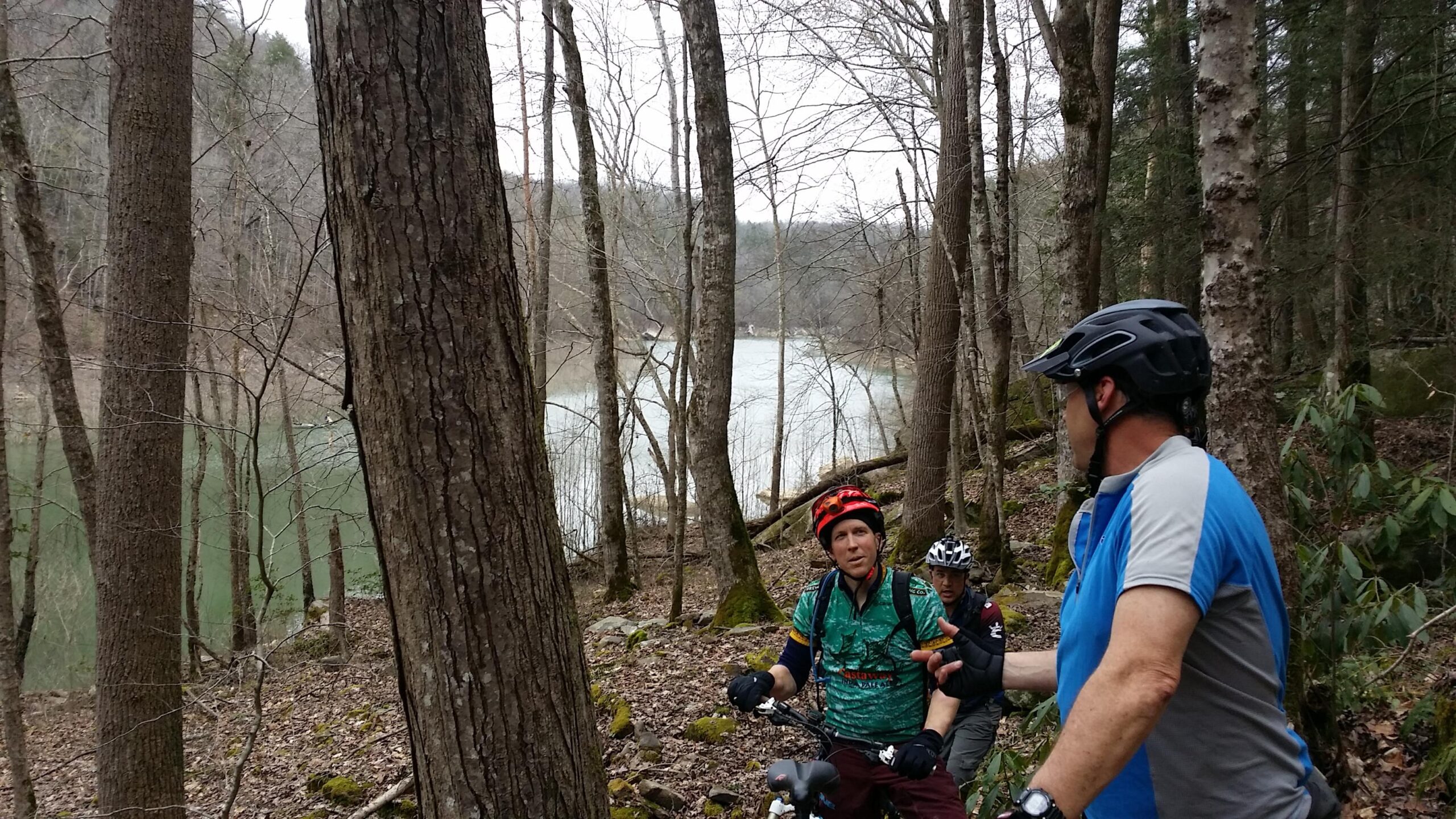 Two mountain bikers pause during a ride in a forested area near a river. The scene shows a mix of trees and rocks, with a glimpse of the water in the background. The bikers are wearing helmets and colorful biking gear, engaged in conversation while surrounded by nature. Laural Lake To Cumberland Falls (sheltowee Trace) mountain bike trail.