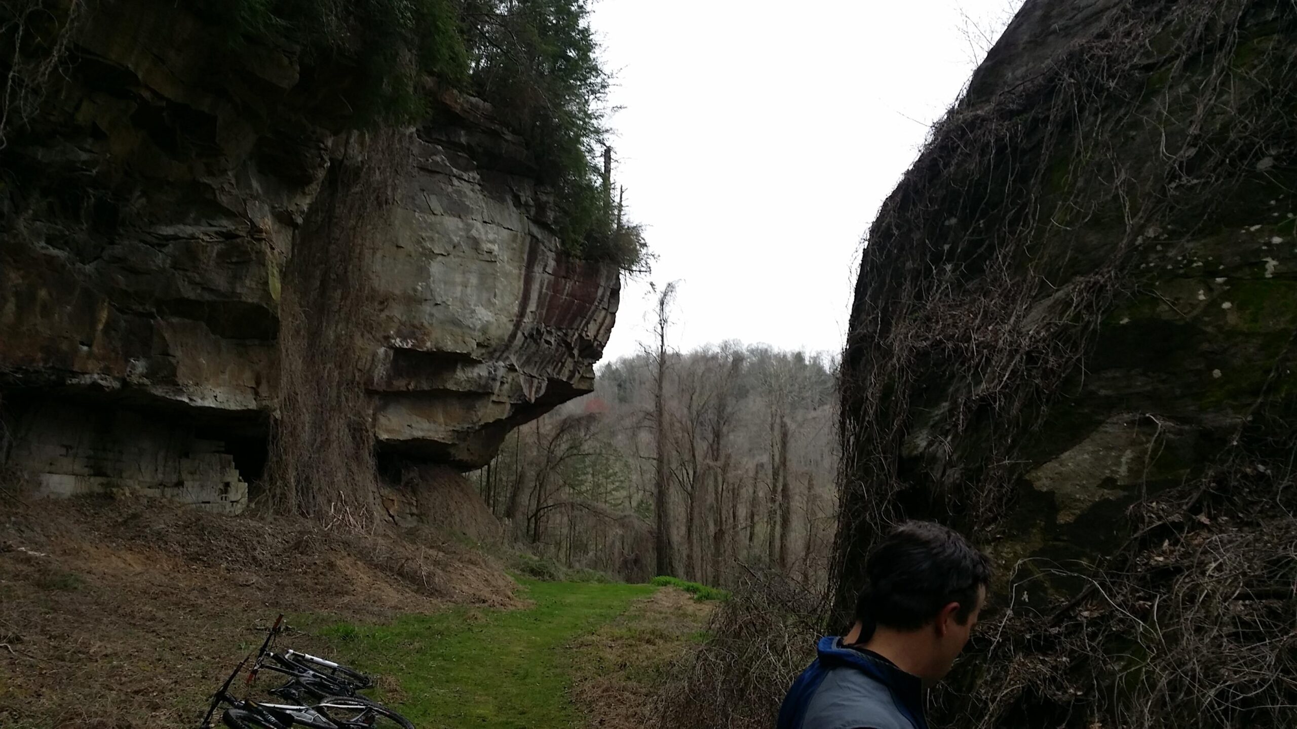 A hiker stands near a rocky outcrop covered in vines, with two mountain bikes leaned against the vegetation. The background features a densely wooded area with leafless trees, and the sky is overcast, suggesting a cool, cloudy day. Laural Lake To Cumberland Falls (sheltowee Trace) mountain bike trail.