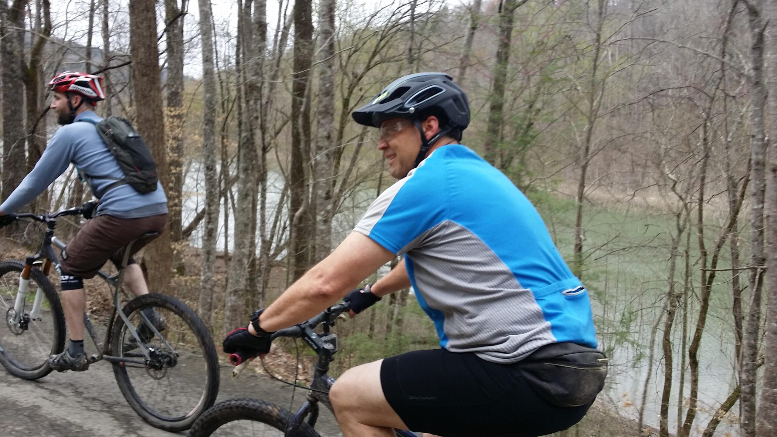 Two cyclists ride their mountain bikes along a dirt path surrounded by trees. One rider, wearing a black helmet and a blue shirt, is in the foreground, while the other, dressed in a gray top and a red helmet, follows behind. In the background, a body of water is visible through the trees. The scene captures the outdoor adventure and natural setting. Laural Lake To Cumberland Falls (sheltowee Trace) mountain bike trail.