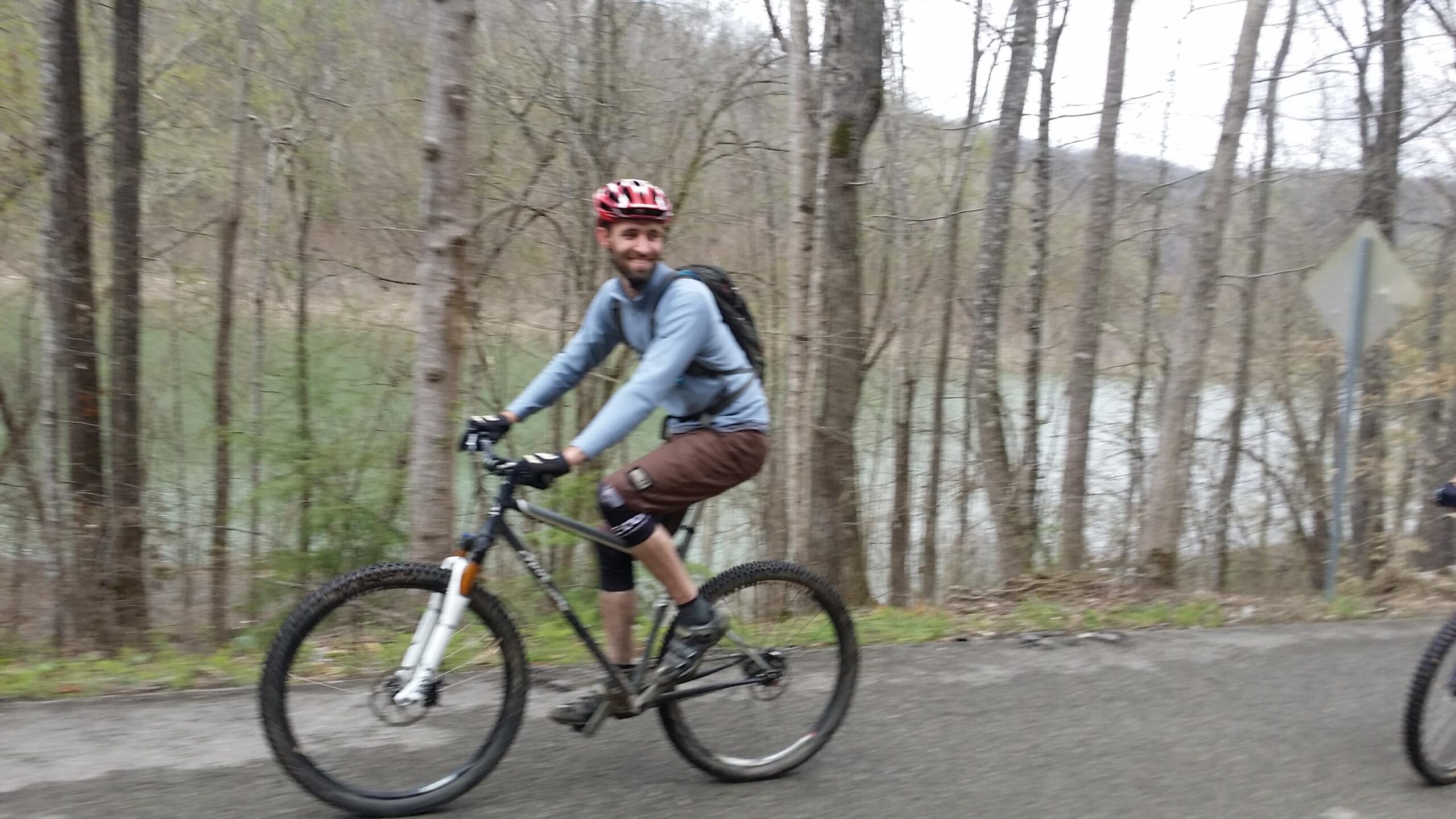 A cyclist riding a mountain bike along a forested path, smiling as he enjoys the outdoors. The background features trees and a body of water, suggesting a scenic trail. Laural Lake To Cumberland Falls (sheltowee Trace) mountain bike trail.