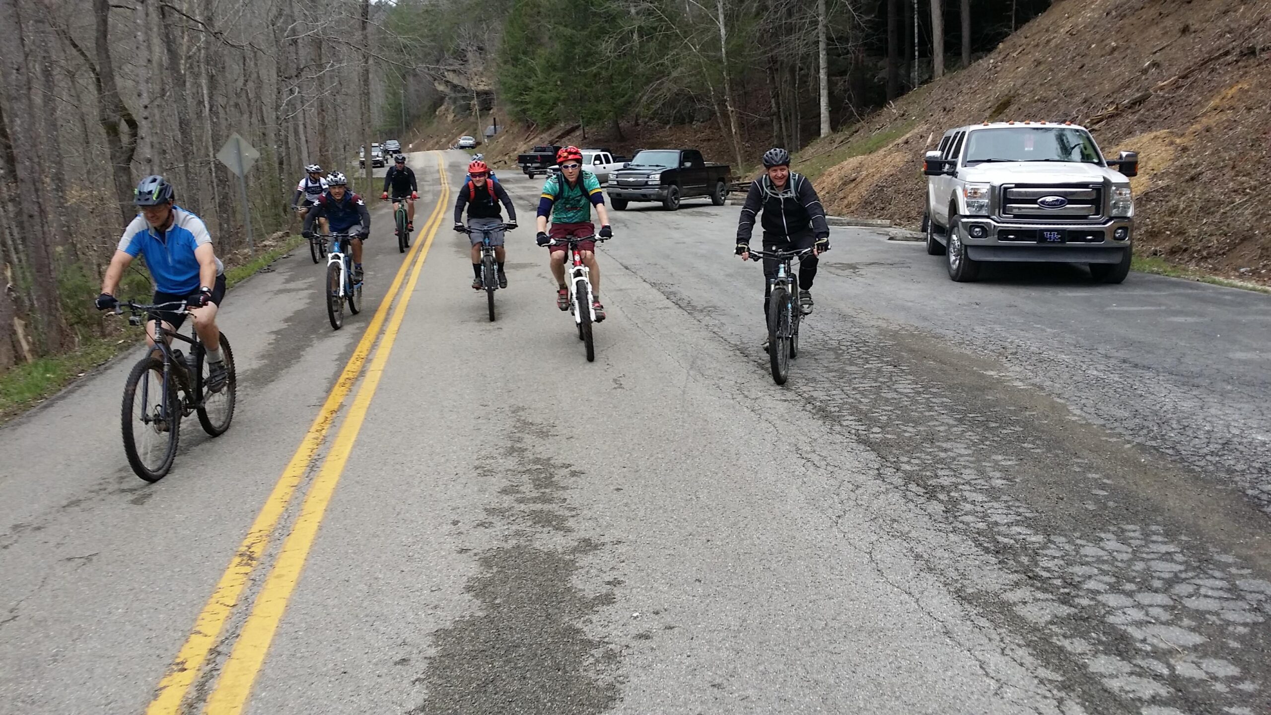 A group of cyclists riding mountain bikes on a paved road lined with trees. Several bikers are actively pedaling, wearing helmets and various cycling attire, while a few vehicles are parked along the roadside. The scene captures a lively outdoor activity in a natural setting. Laural Lake To Cumberland Falls (sheltowee Trace) mountain bike trail.