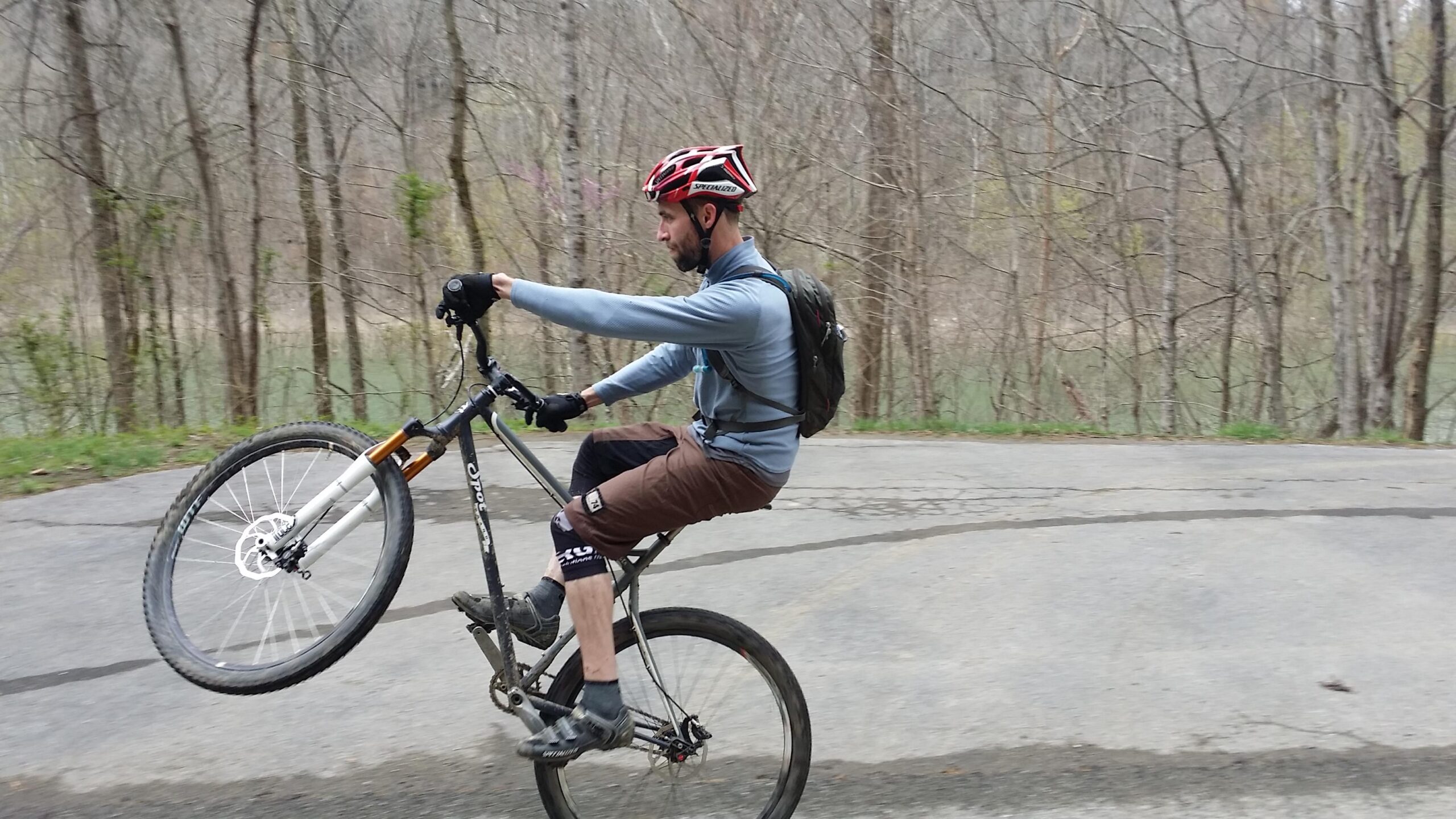 A person riding a mountain bike on a paved path, performing a wheelie. The rider is wearing a helmet and gloves, dressed in a long-sleeve shirt and shorts. Surrounding them are bare trees and a view of a body of water in the background, indicating a natural setting. Laural Lake To Cumberland Falls (sheltowee Trace) mountain bike trail.