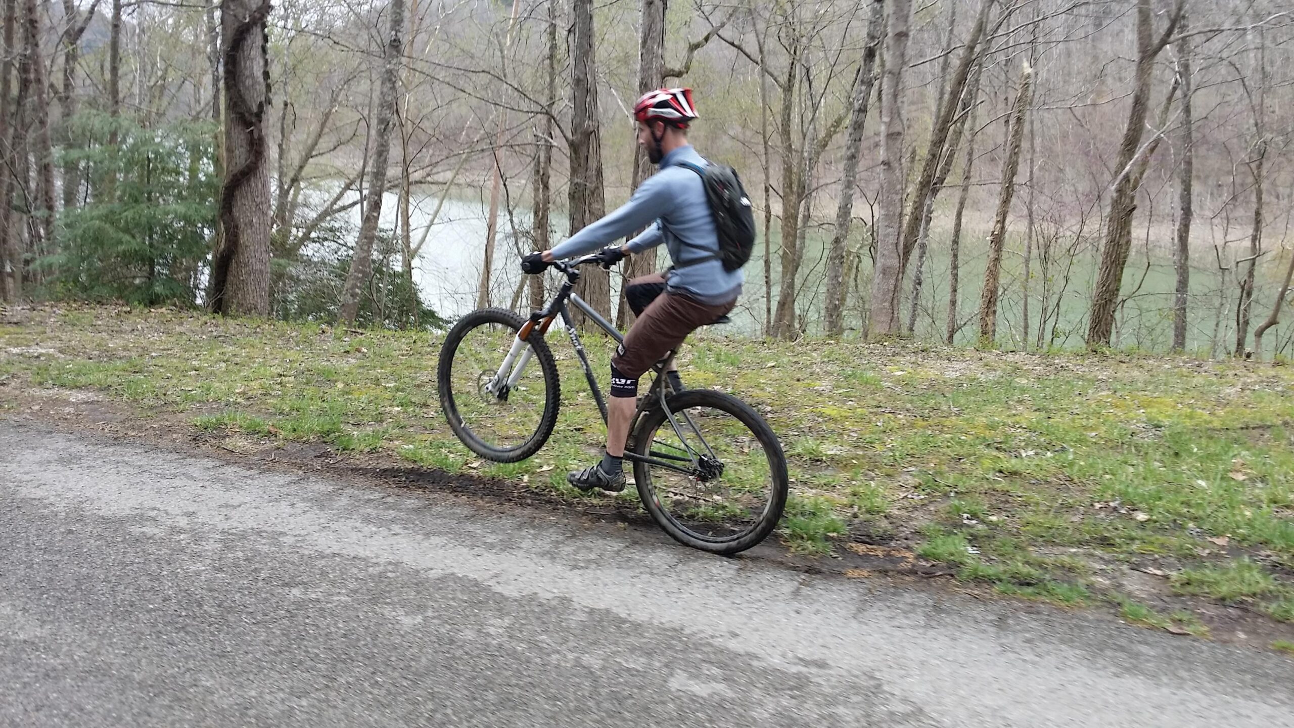 A person riding a mountain bike on one wheel along a path next to a lake, surrounded by trees and greenery. The cyclist is wearing a helmet and casual clothing, demonstrating a stunt while enjoying the outdoors. Laural Lake To Cumberland Falls (sheltowee Trace) mountain bike trail.