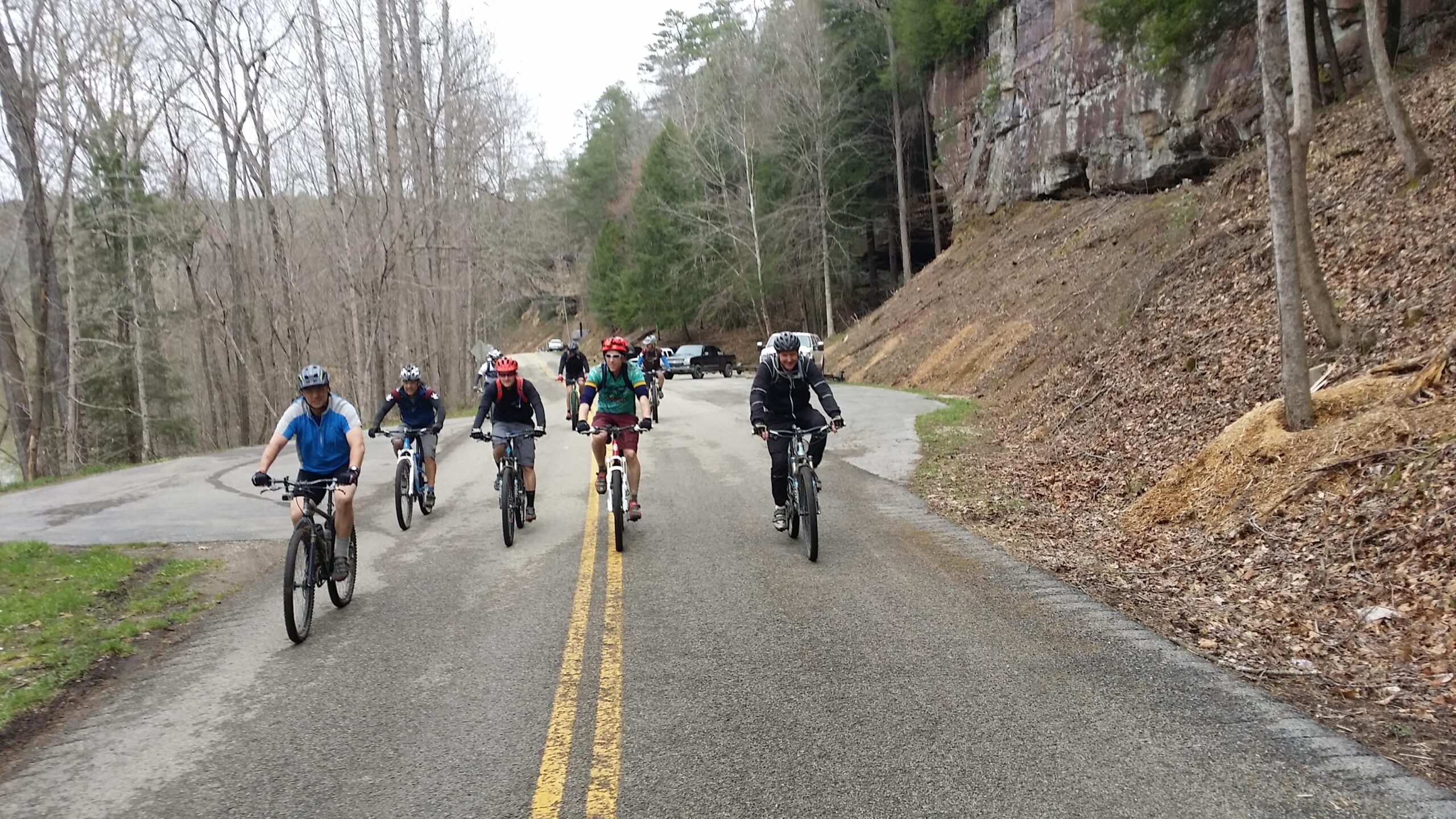 A group of eight mountain bikers riding on a winding road surrounded by bare trees and rocky terrain. Some bikers wear helmets and sporty attire, while a couple of parked vehicles are visible in the background. The scene captures a sunny day in a natural setting, highlighting an outdoor cycling adventure. Laural Lake To Cumberland Falls (sheltowee Trace) mountain bike trail.