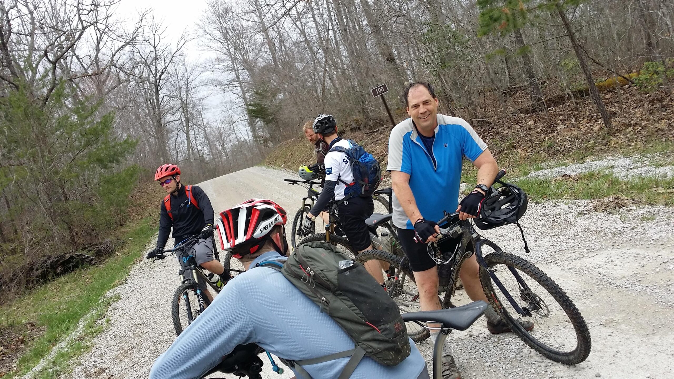 Four mountain bikers pause on a gravel road surrounded by trees. One rider in a blue shirt smiles at the camera, while others are engaged in conversation or adjusting their gear. The scene captures a moment of camaraderie during an outdoor biking adventure. Laural Lake To Cumberland Falls (sheltowee Trace) mountain bike trail.