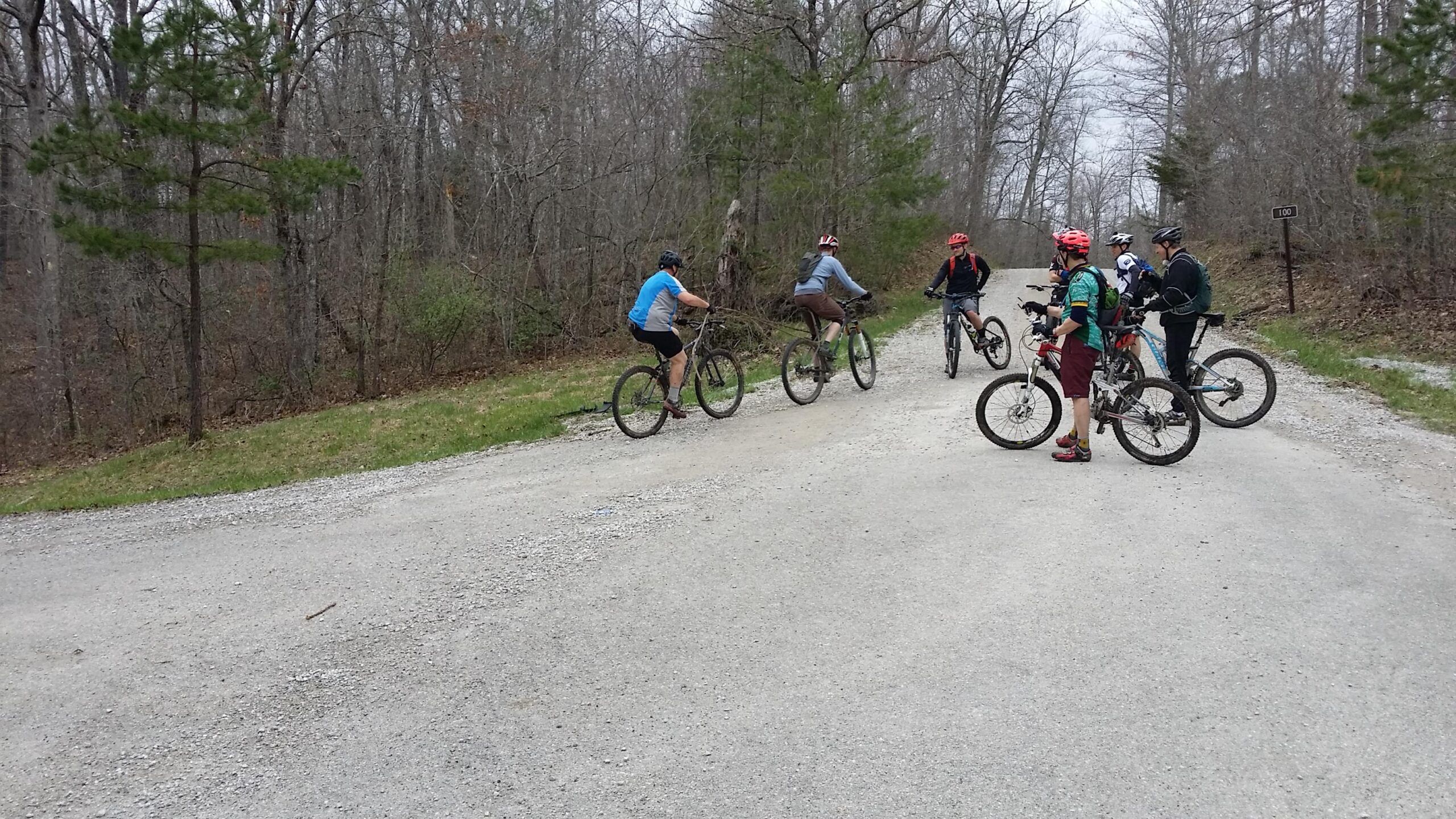 A group of six mountain bikers is gathered at a fork in a gravel trail surrounded by trees, with some wearing helmets and colorful cycling attire. The atmosphere is overcast, suggesting a cool, outdoor setting. Sheltowee Trace - Laurel Lake Trail mountain bike trail.