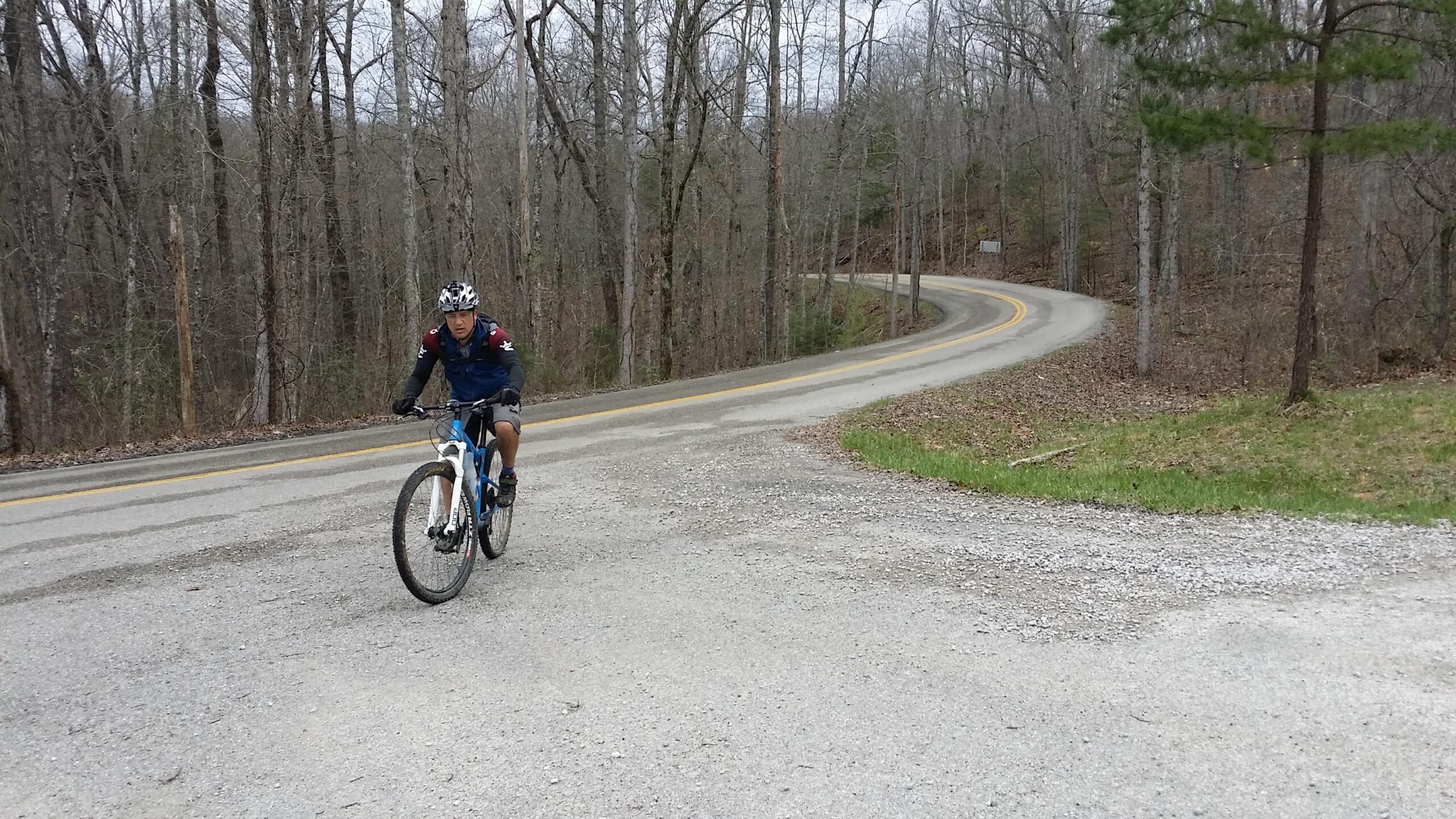 A cyclist riding a blue mountain bike on a gravel road with trees in a partly wooded area. The path winds to the right, and the cyclist is approaching a fork in the road, surrounded by bare trees and some greenery. The sky is overcast. Sheltowee Trace - Laurel Lake Trail mountain bike trail.