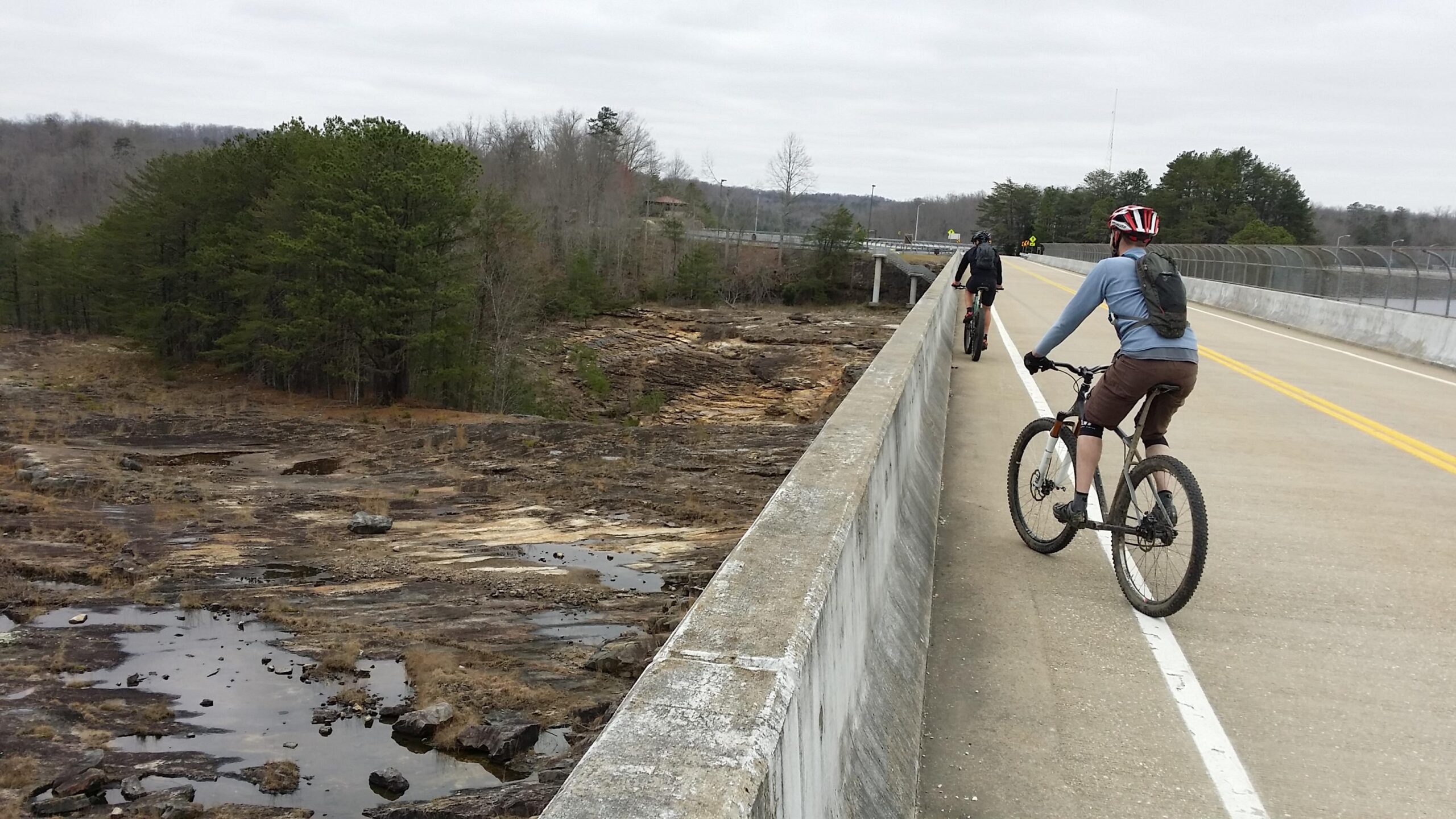 Two mountain bikers ride along a bridge with a concrete surface and yellow lane markings, surrounded by a rocky landscape and sparse trees. The sky is overcast, suggesting cool weather. One biker is wearing a helmet and a backpack, while the other is in cycling attire, both enjoying their ride on the elevated path. Sheltowee Trace - Laurel Lake Trail mountain bike trail.