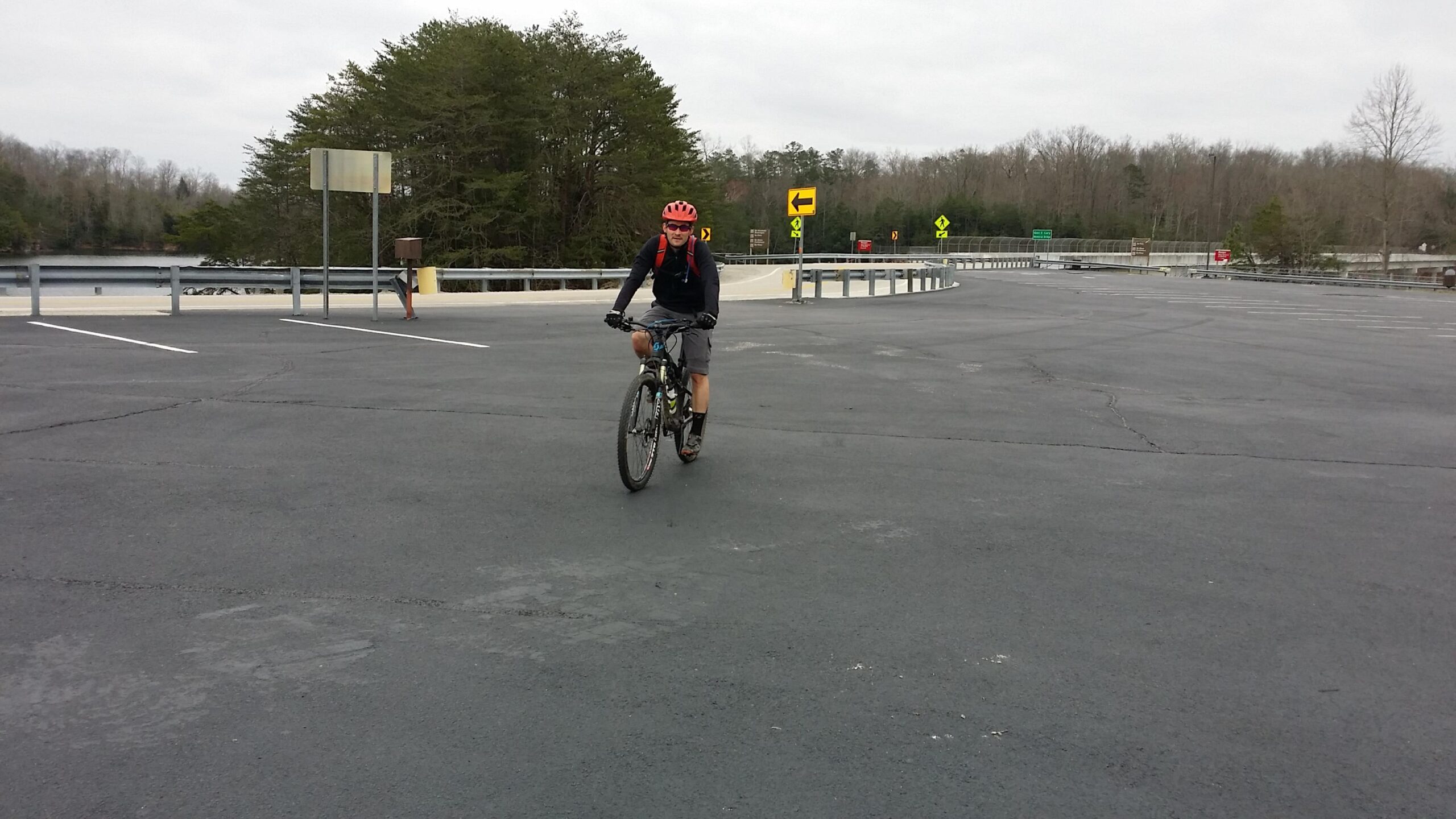 A cyclist wearing a red helmet and backpack rides a mountain bike on an empty asphalt parking lot, with trees and a body of water visible in the background under a cloudy sky. Road signs can be seen in the distance. Sheltowee Trace - Laurel Lake Trail mountain bike trail.