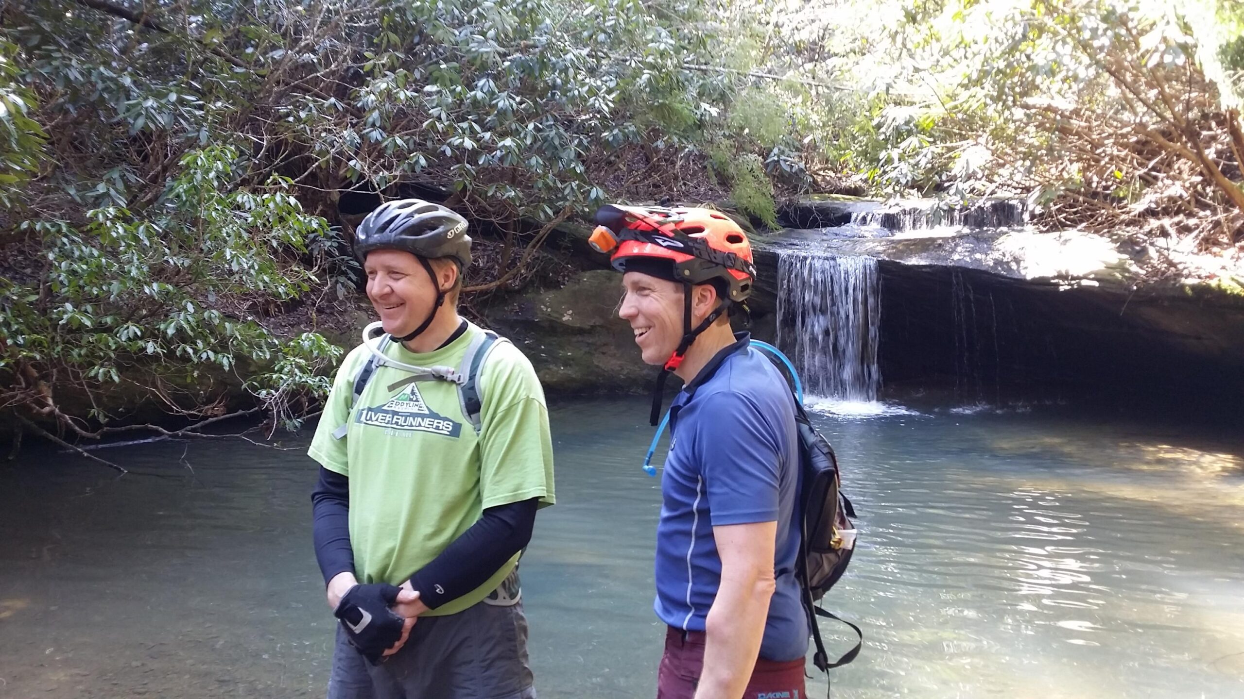 Two men are standing by a tranquil creek in a forested area, smiling and enjoying the scenery. One man, wearing a light green shirt and a black bicycle helmet, has his hands clasped in front of him. The other man, dressed in a blue shirt with a red helmet equipped with a light, has a backpack on and is laughing. In the background, a small waterfall cascades into the creek, surrounded by lush greenery. Cane Creek (sheltowee Trace Trail) mountain bike trail.