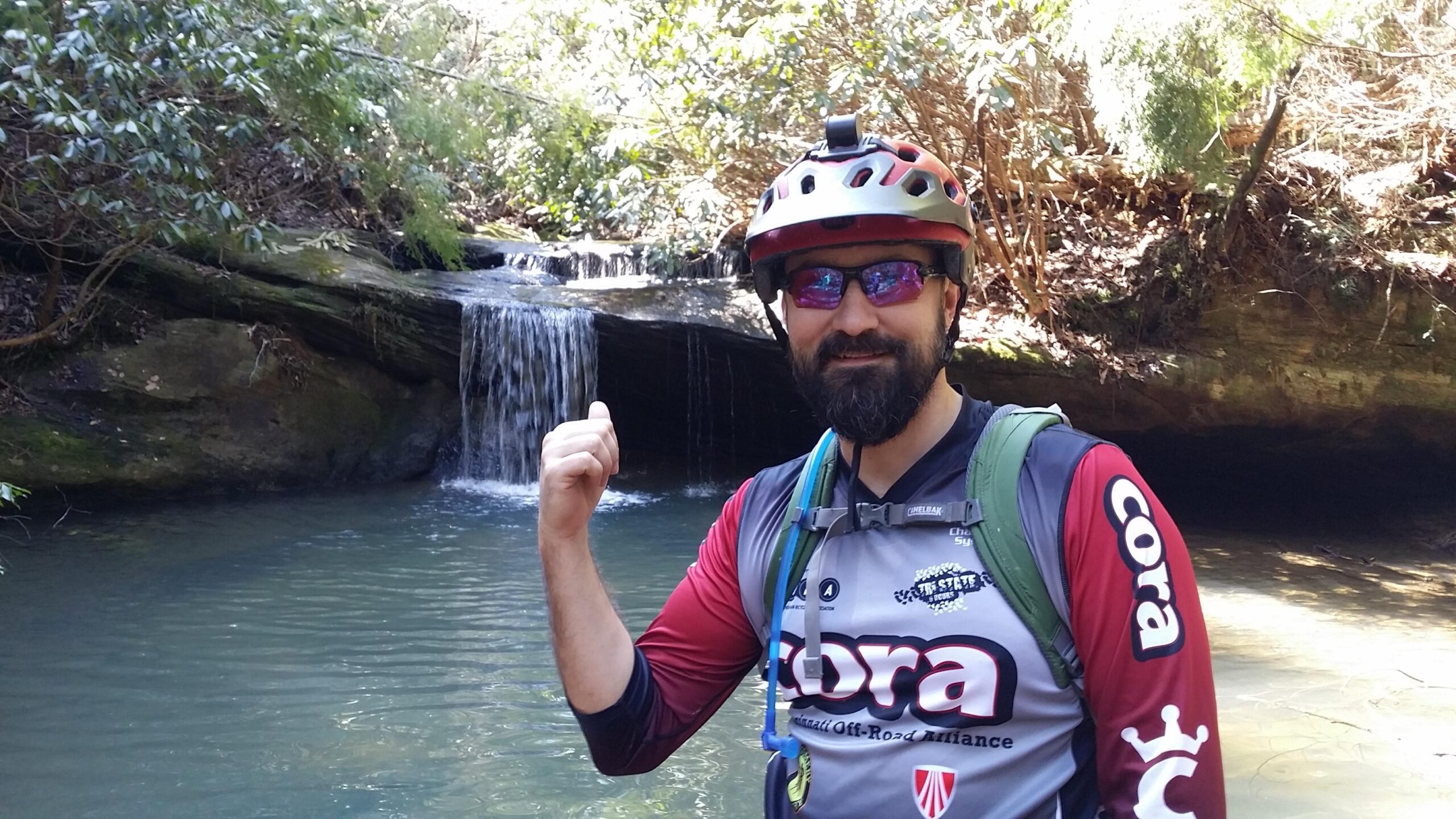 A person wearing a helmet and sunglasses stands by a small waterfall and a tranquil pool of water, giving a thumbs-up. The scene is surrounded by lush greenery, indicating a natural outdoor setting. The individual is dressed in a colorful athletic shirt, suitable for biking or outdoor activities. Cane Creek (sheltowee Trace Trail) mountain bike trail.