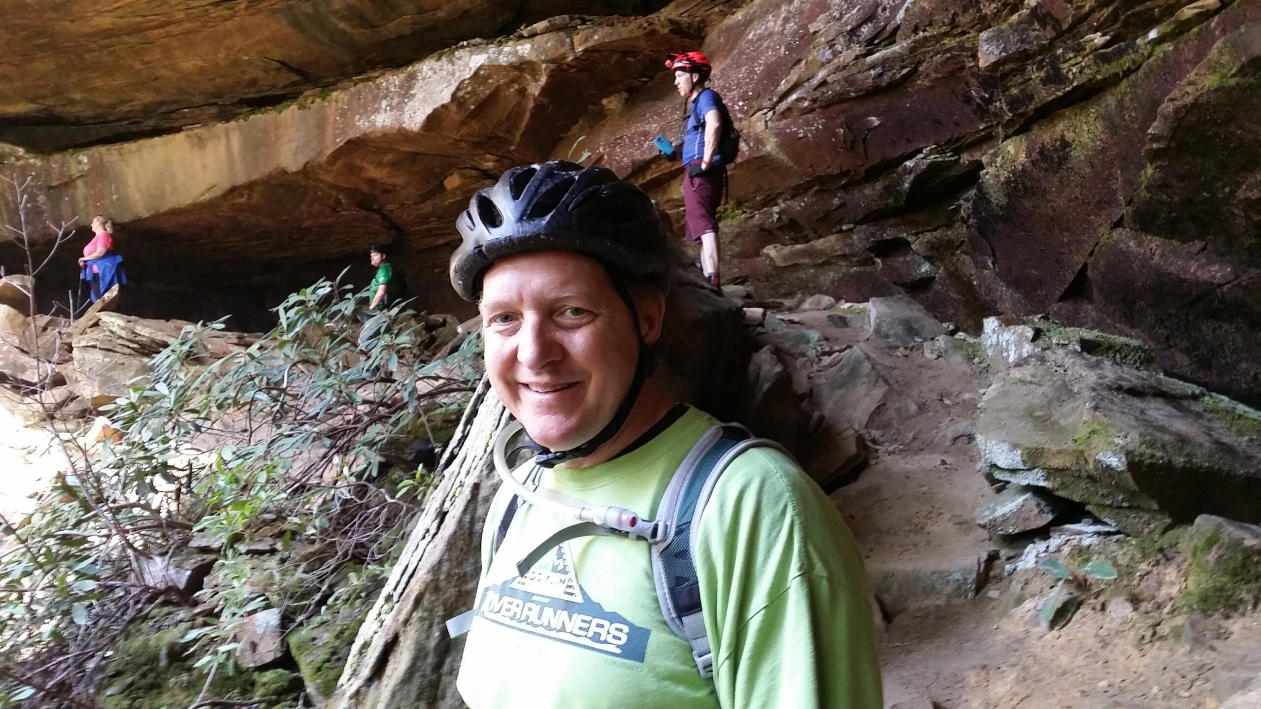 A man wearing a green t-shirt and a bike helmet smiles at the camera while standing on a rocky path. In the background, two people explore a cave-like area, with one person standing on a ledge and another further back. Lush green foliage surrounds the scene, set against the textured rock formations of the cave. Cane Creek (sheltowee Trace Trail) mountain bike trail.