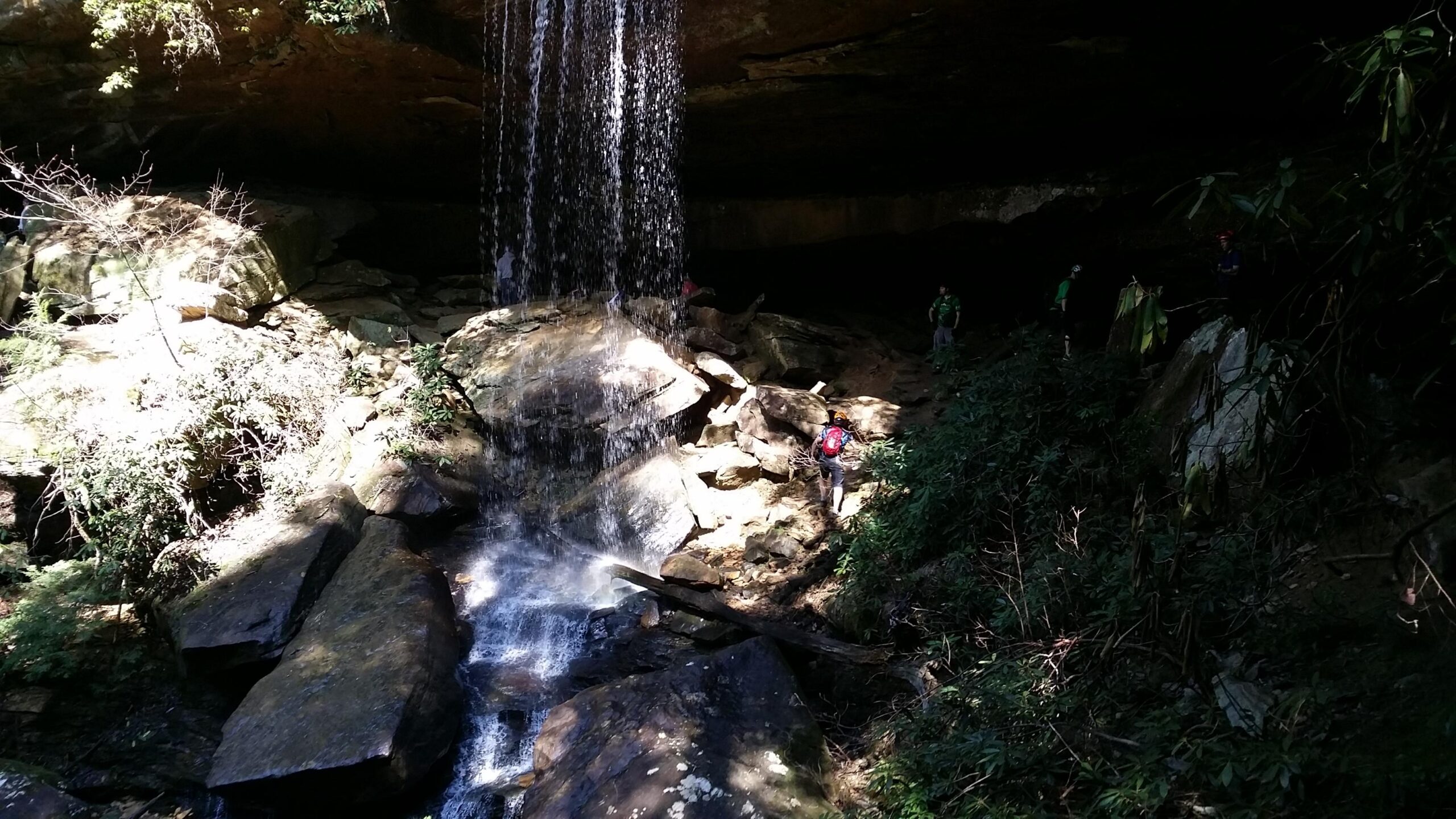 A waterfall cascading over rocks into a serene area surrounded by lush greenery and moss-covered stones. Several hikers can be seen exploring the rocky terrain beneath the waterfall, with sunlight filtering through the trees above. Cane Creek (sheltowee Trace Trail) mountain bike trail.