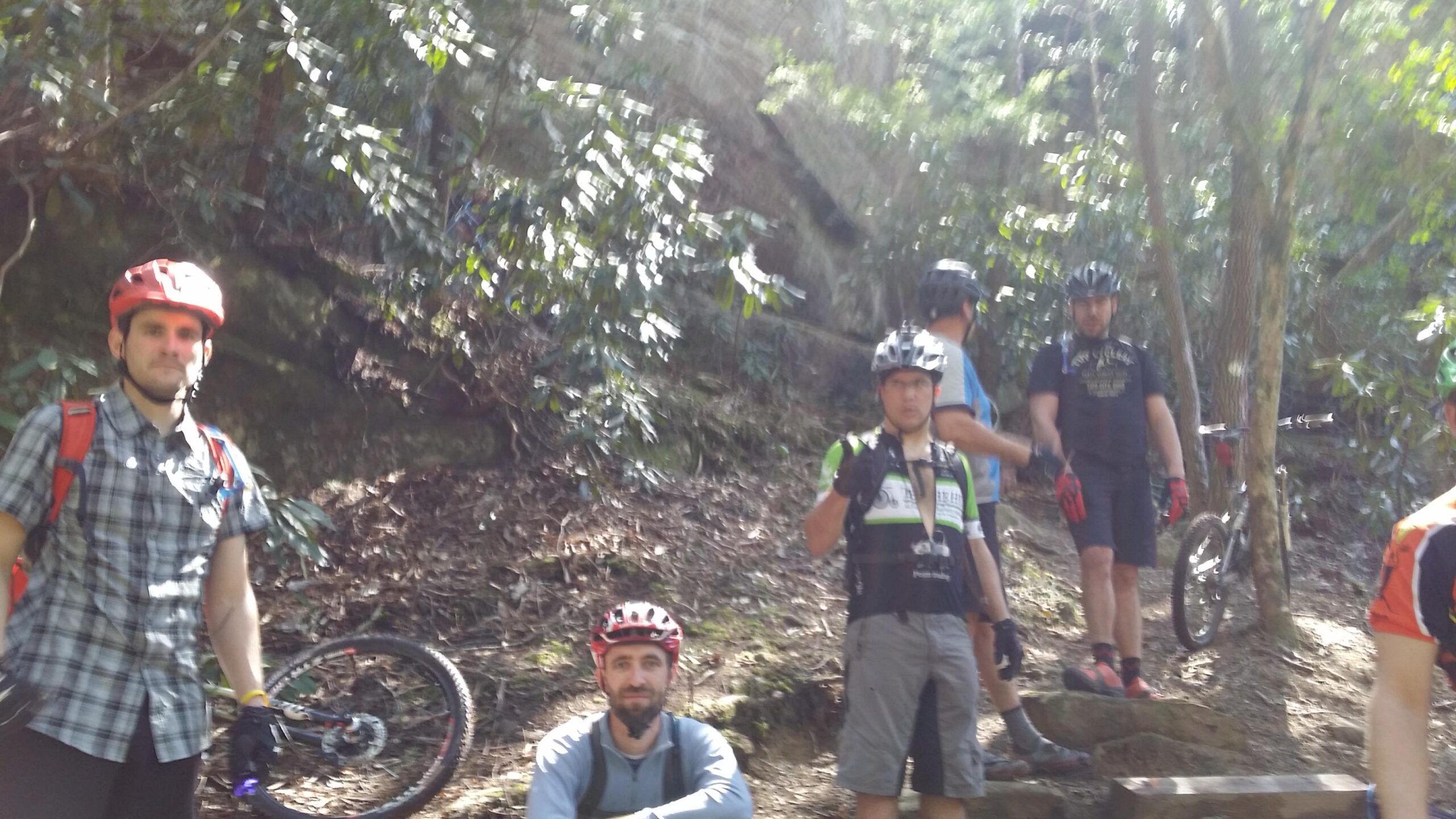 A group of six mountain bikers, dressed in casual biking attire and helmets, are gathered on a dirt path surrounded by lush greenery. Some are posing for the camera, while others are engaged in conversation. Mountain bikes are parked nearby, with the sunlight filtering through the trees, creating a lively outdoor atmosphere. Cane Creek (sheltowee Trace Trail) mountain bike trail.