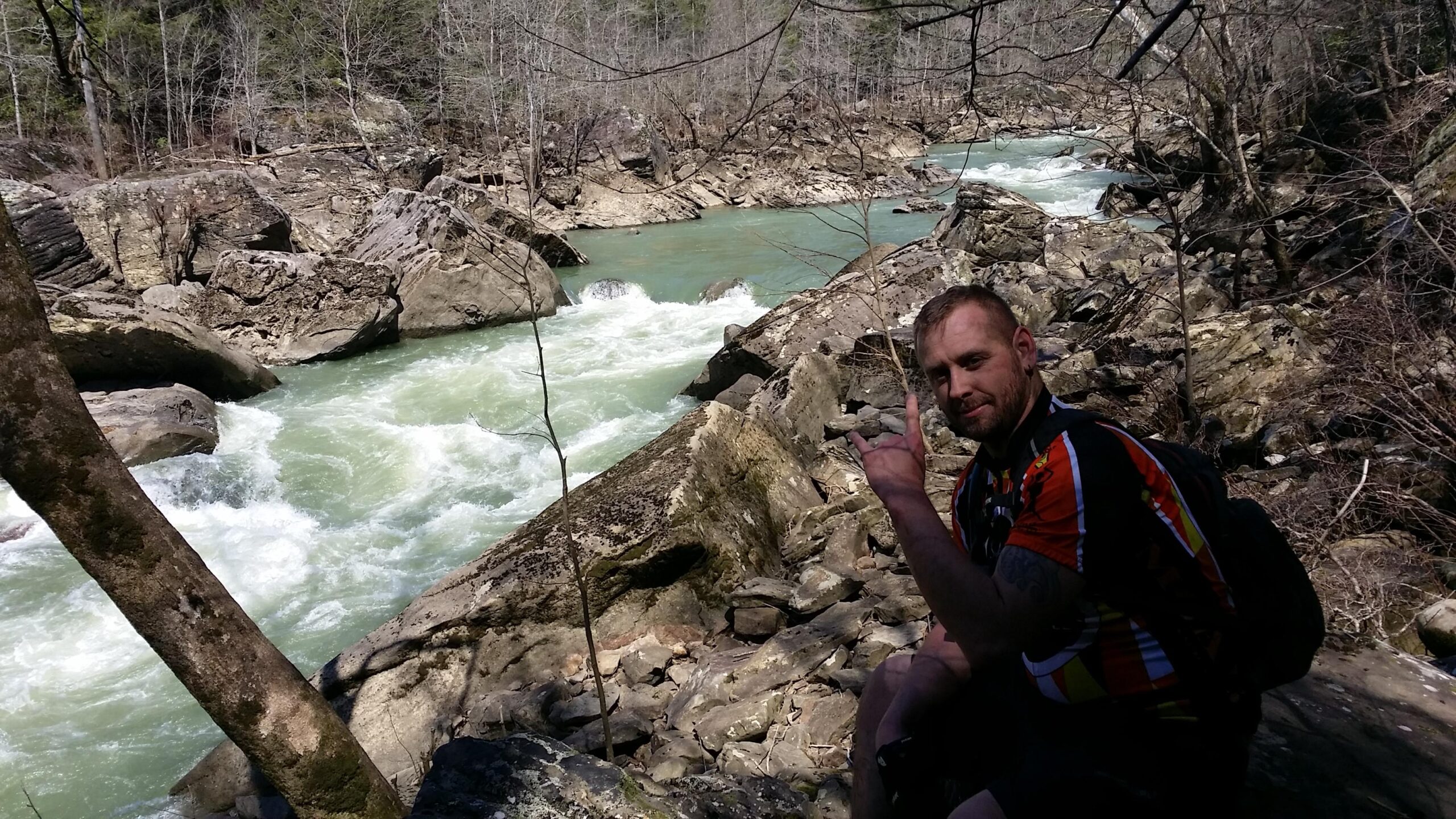A man sitting on rocks by a rushing river, making a hand gesture. The scene features rocky terrain and trees in the background, with clear, flowing water in shades of green and white. The weather is sunny, indicating a pleasant outdoor environment. Cane Creek (sheltowee Trace Trail) mountain bike trail.