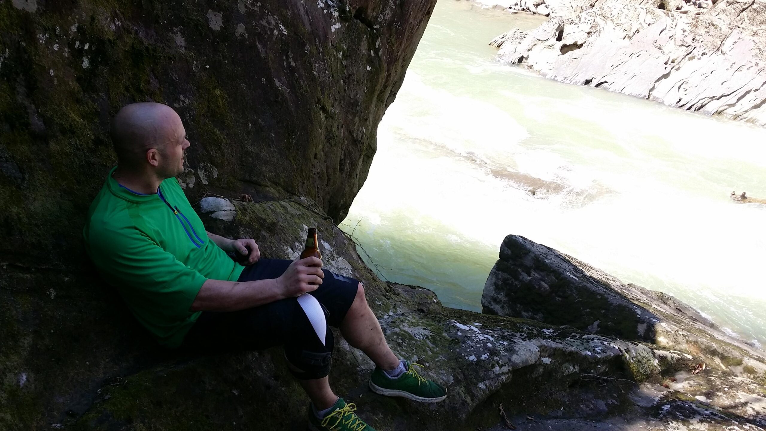 A person wearing a green shirt and black shorts sits on a rock near a river. They are holding a bottle and looking thoughtfully at the water, which features swift currents. The surrounding area includes large stones and moss-covered surfaces, suggesting a natural outdoor setting. Cane Creek (sheltowee Trace Trail) mountain bike trail.
