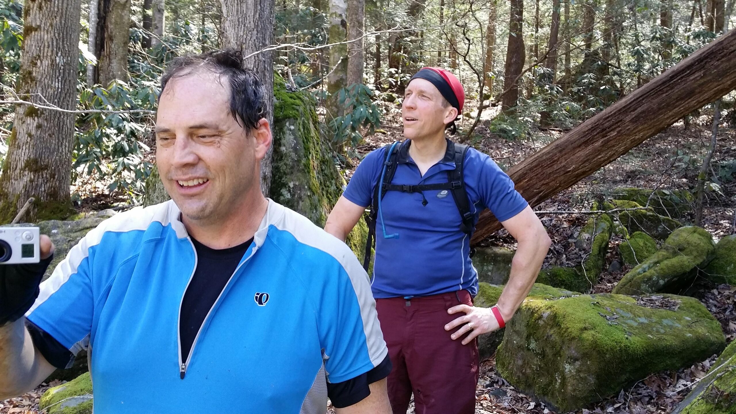 Two men in athletic clothing are standing in a wooded area. One man is smiling and holding a camera, while the other appears to be looking off into the distance. Surrounding them are trees and moss-covered rocks, indicating a hiking or outdoor setting. The sunlight filters through the foliage, creating a lively atmosphere. Cane Creek (sheltowee Trace Trail) mountain bike trail.