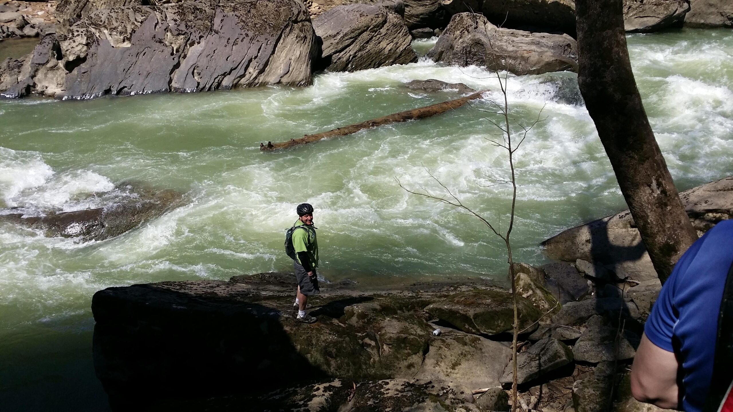 A person wearing a green shirt and a helmet stands on a rocky riverbank, looking out at a rushing river. The water is a light greenish color, with white rapids and a log floating in the current. Surrounding the river are large rocks and trees, creating a natural landscape. The scene is bright and sunny, suggesting a warm day outdoors. Cane Creek (sheltowee Trace Trail) mountain bike trail.