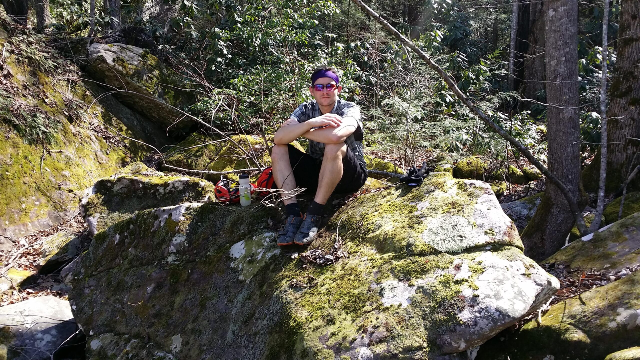 A person sitting on a moss-covered rock in a forested area, wearing a plaid shirt, shorts, and sunglasses. Nearby, there is a water bottle and a red backpack. The background features trees and greenery, typical of a natural outdoor setting. Cane Creek (sheltowee Trace Trail) mountain bike trail.