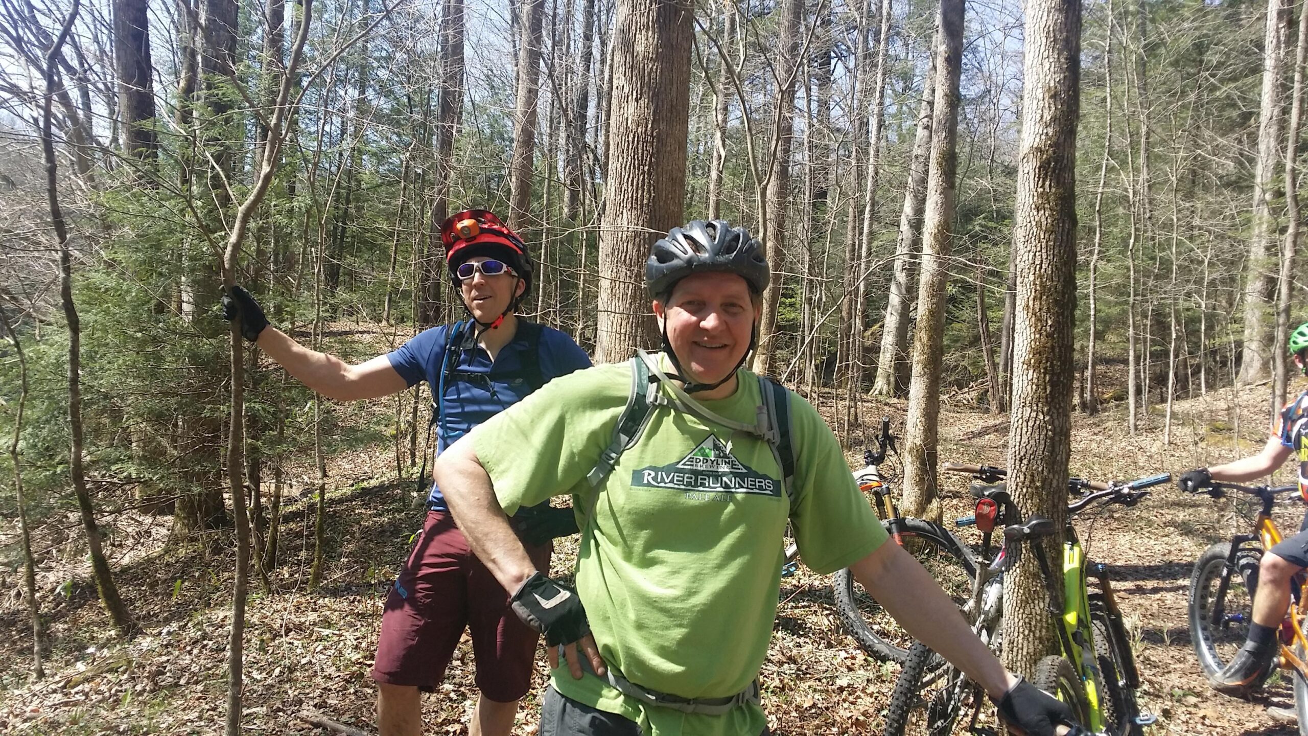 Two men are standing in a wooded area, surrounded by trees and foliage. One man, wearing a green shirt and gloves, is smiling and has a hand on his hip. The second man, with a helmet and sunglasses, is leaning on a small tree, dressed in a blue shirt and maroon shorts. In the background, several mountain bikes are parked. It's a sunny day, suggesting an outdoor recreational activity, possibly mountain biking. Cane Creek (sheltowee Trace Trail) mountain bike trail.