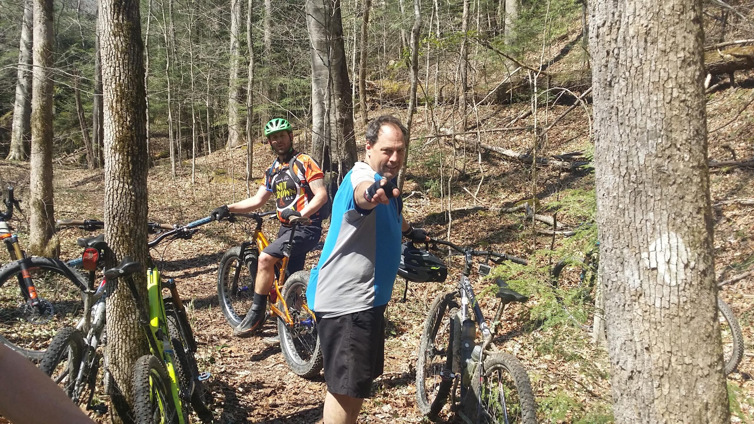 A group of mountain bikers stopped on a forest trail, with two men in the foreground. One man in a blue and gray shirt is pointing at the camera, while the other, wearing an orange and black jersey, stands beside a mountain bike. The surrounding area features trees, underbrush, and fallen leaves, indicating an outdoor adventure in a wooded environment. Cane Creek (sheltowee Trace Trail) mountain bike trail.