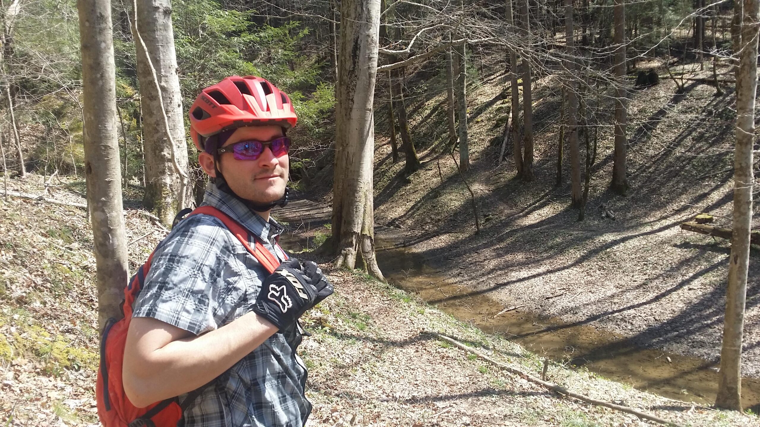 A person wearing a red helmet, sunglasses, and gloves stands on a wooded trail beside a small stream. They are wearing a plaid shirt and a backpack, with trees and fallen leaves visible in the background, indicating a sunny day in a forested area. Cane Creek (sheltowee Trace Trail) mountain bike trail.