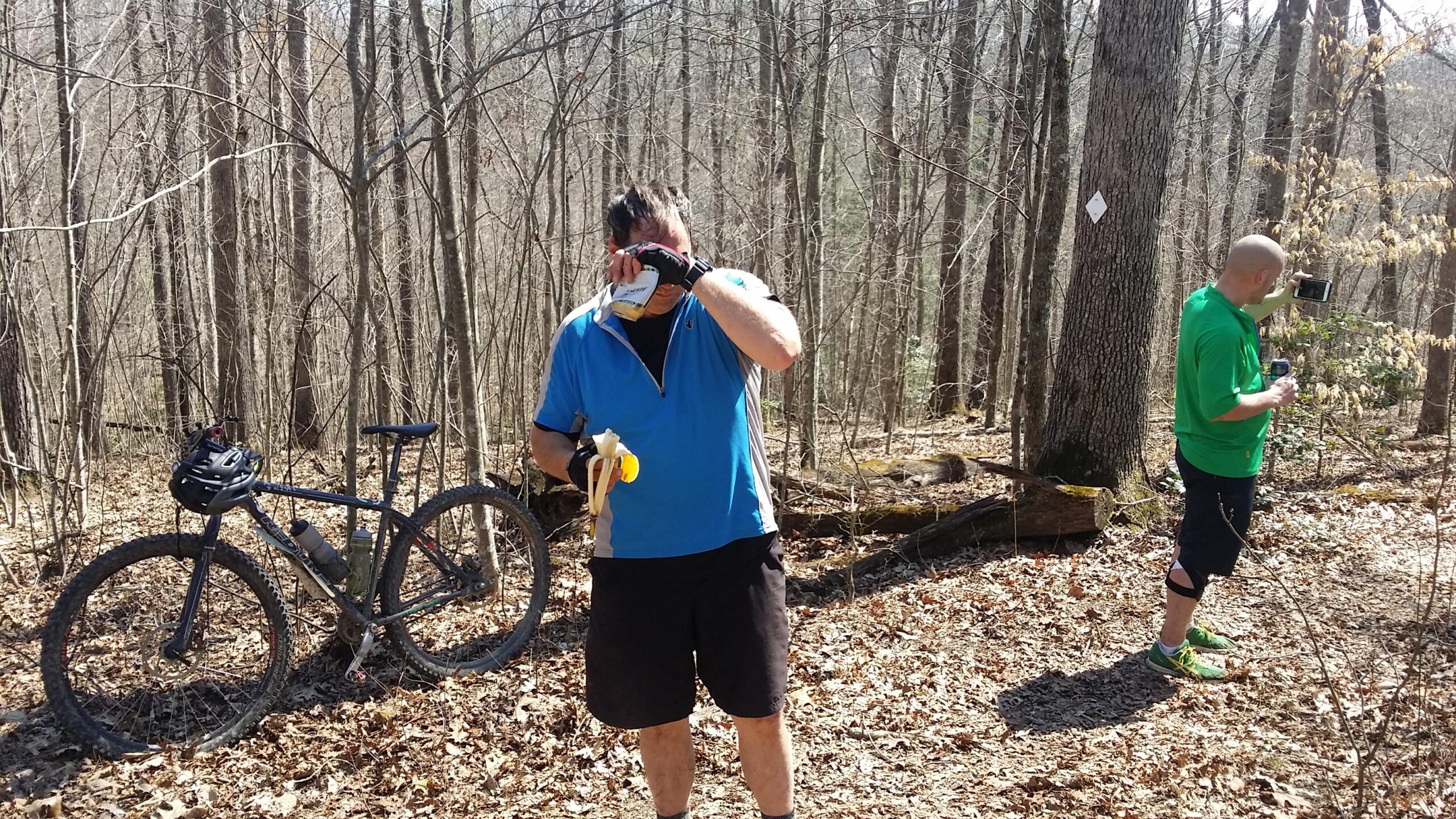 Two men in a wooded area are taking a break during a mountain biking trip. One man, wearing a blue shirt and black shorts, is holding a banana and drinking from a can. The other man, dressed in a green shirt and black shorts, is taking a photo with his phone. A mountain bike is parked nearby, and the surroundings have bare trees and fallen leaves. Cane Creek (sheltowee Trace Trail) mountain bike trail.