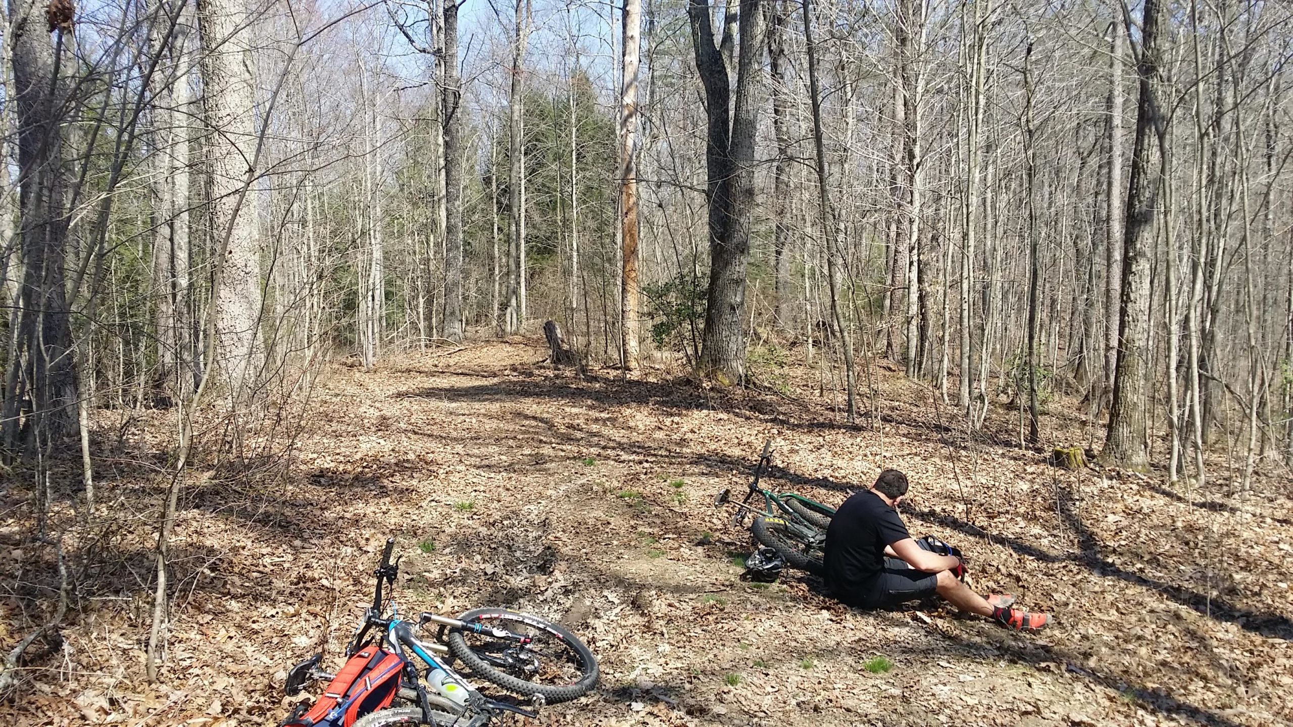 A mountain biker sits on the forest floor, resting near two parked bicycles, amidst a wooded area with bare trees and scattered leaves on the ground. The path curves into the distance, suggesting a trail in a natural outdoor setting. Cane Creek (sheltowee Trace Trail) mountain bike trail.