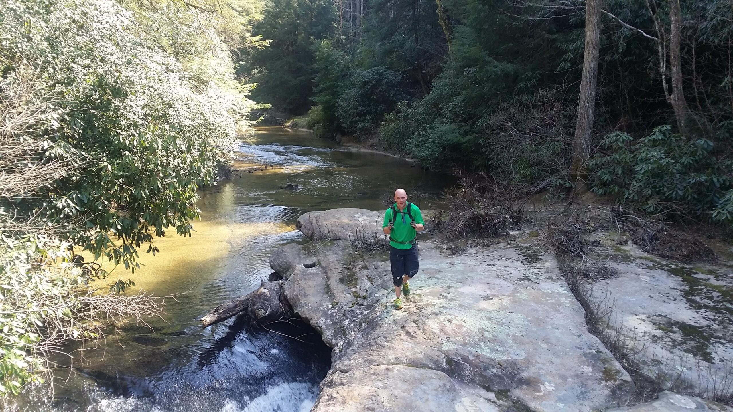 A person with a backpack is walking along a rocky riverbank surrounded by greenery. The water flows gently beside them, reflecting the sunlight, while lush trees and shrubs line the area. Cane Creek (sheltowee Trace Trail) mountain bike trail.