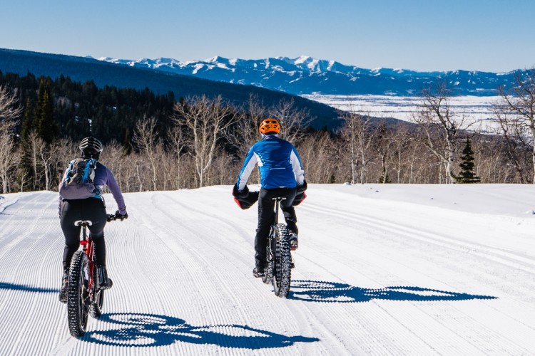 Two cyclists ride on a snowy, groomed trail with a stunning mountainous landscape in the background. The rider on the left, wearing a backpack, has a black bicycle, while the one on the right is in a bright orange helmet and blue jacket, biking on a thicker-tired bike designed for snow. The scene captures a clear, sunny day with a blue sky.