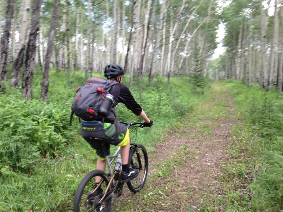 A person wearing a helmet and backpack is riding a mountain bike along a narrow dirt path in a wooded area. The scene is surrounded by tall trees with green foliage, creating a peaceful, natural setting. Snake Indian Falls mountain bike trail.