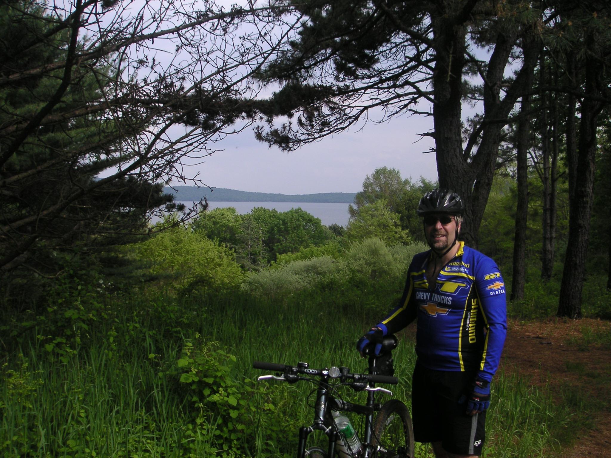 A mountain biker in a blue and yellow jersey stands next to his bicycle on a wooded trail, with a lake visible in the background. The scene features lush greenery and tall trees, creating a serene outdoor atmosphere. Round Valley Recreation Area mountain bike trail.