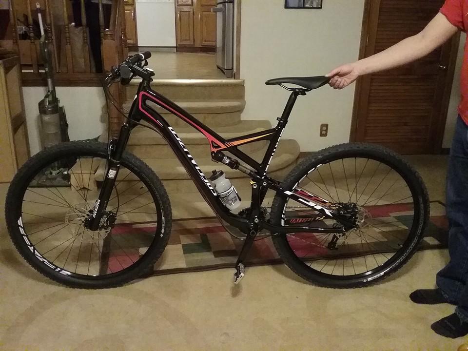 Specialized Camber Comp 29: A mountain bike with a black and orange frame is positioned on a carpeted floor. The bike features thick tires, a water bottle mounted on the frame, and a person’s hand holding the seat. In the background, there is a staircase leading to a kitchen area.