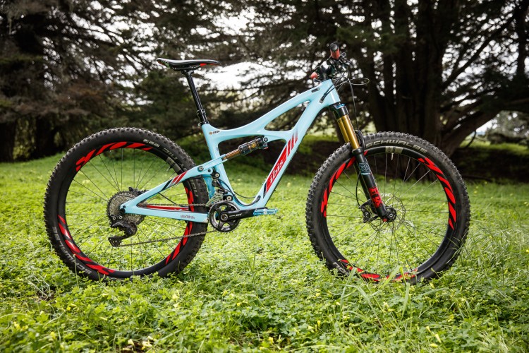 A mountain bike with a light blue frame and black wheels featuring red accents, standing on a grassy area with trees in the background. The bike is equipped with a suspension system and has a sleek design, showcasing its readiness for outdoor trails.