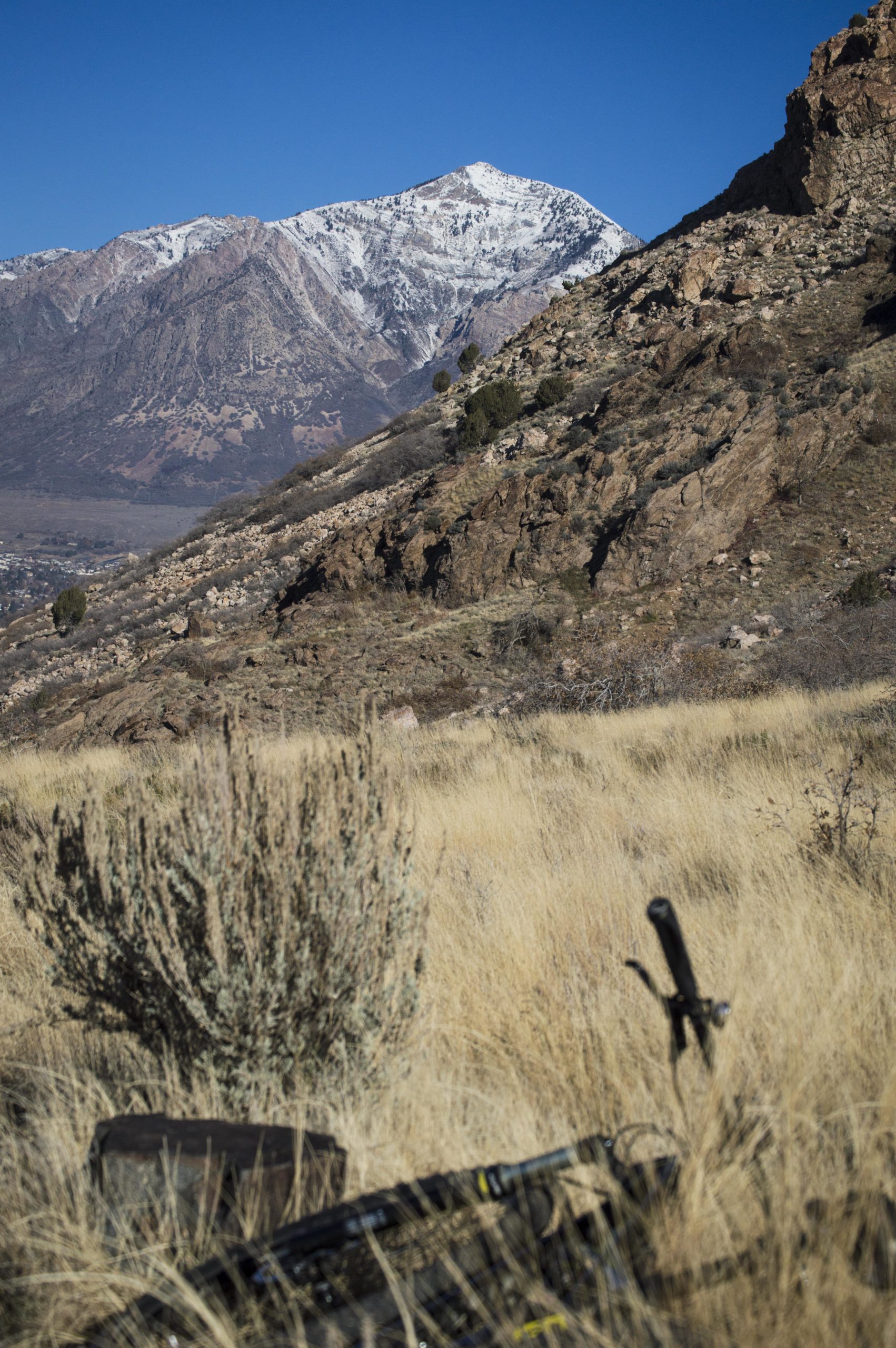 A scenic view of a mountainous landscape featuring snow-capped peaks in the distance, with rocky terrain and dry, tall grass in the foreground. The sky is clear and blue, enhancing the rugged beauty of the natural setting. A blurred outline of a rock and some equipment is visible in the foreground. Bonneville Shoreline Trail - Ogden Section mountain bike trail.