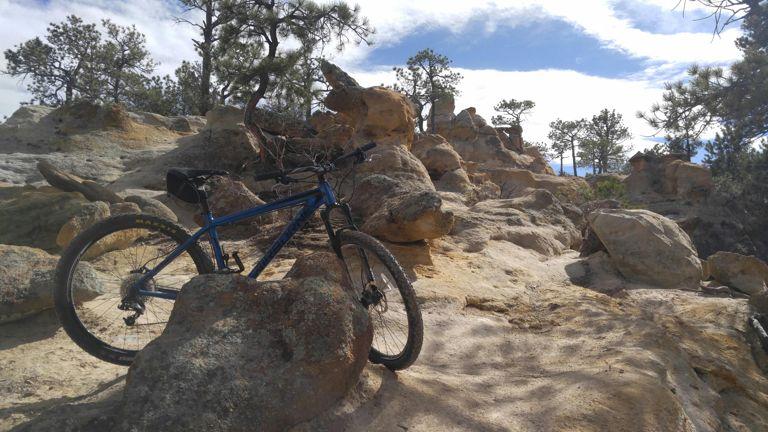 A blue mountain bike is resting against a large rock amid a rugged, rocky landscape, with scattered boulders and pine trees in the background under a partly cloudy sky. Palmer Park mountain bike trail.