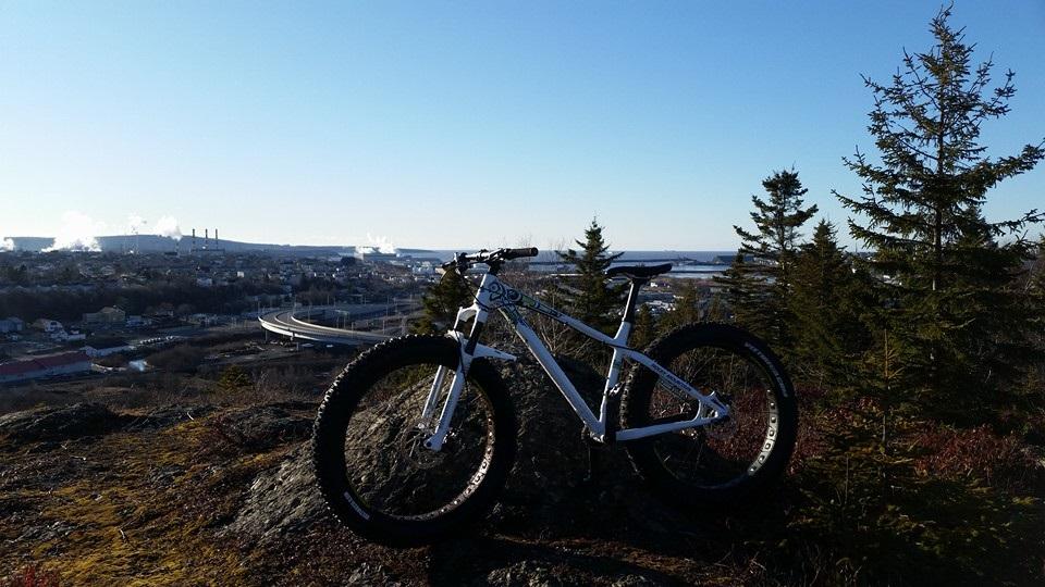 A white mountain bike is positioned on a rocky outcrop, overlooking a scenic view of a town with smokestacks in the distance. The landscape features trees and a clear blue sky, indicating a bright day. The bike’s large tires and sturdy frame suggest it is designed for rugged terrain.